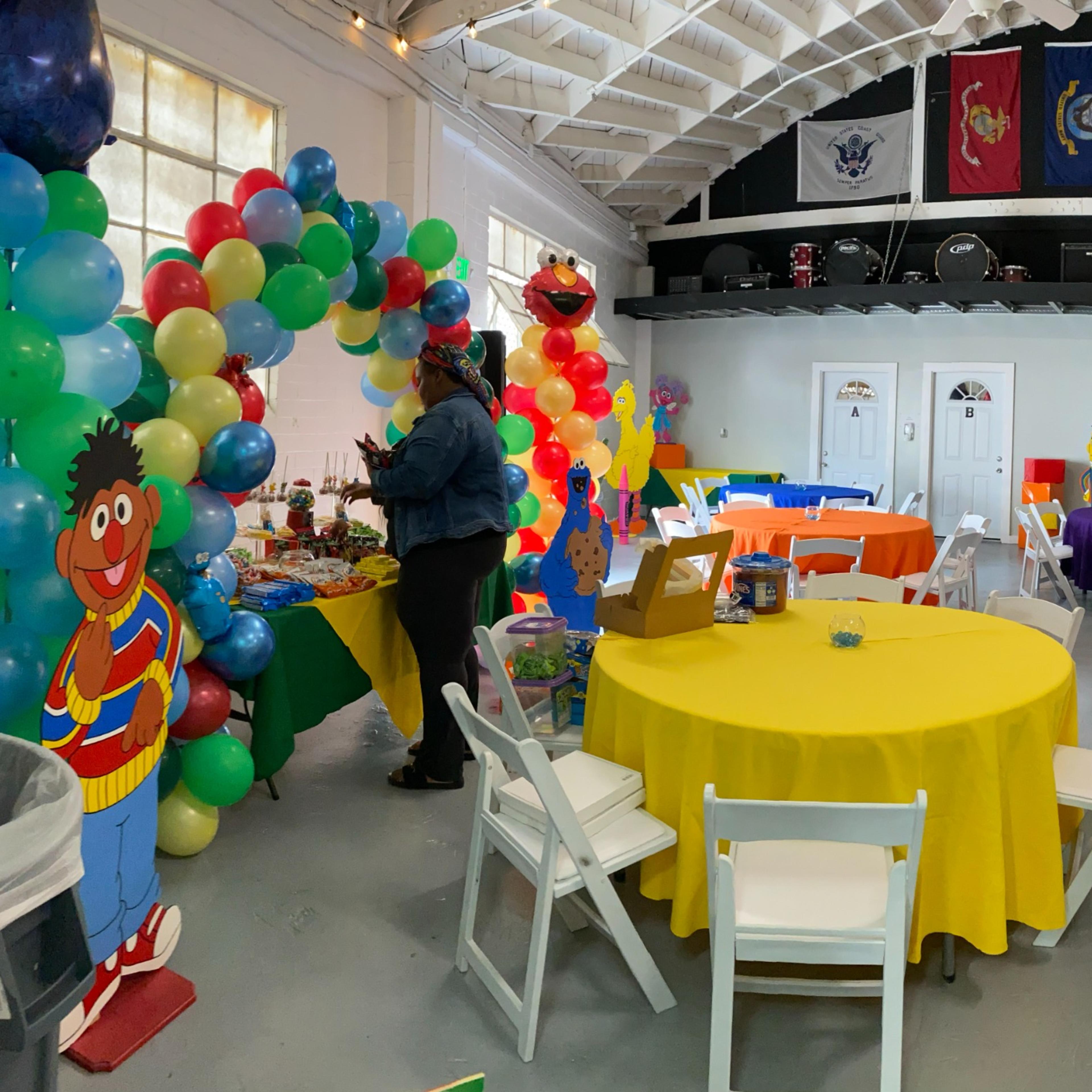 A festive indoor party space decorated with colorful tables, balloons, and a large inflatable structure, with various themed decorations along the walls.