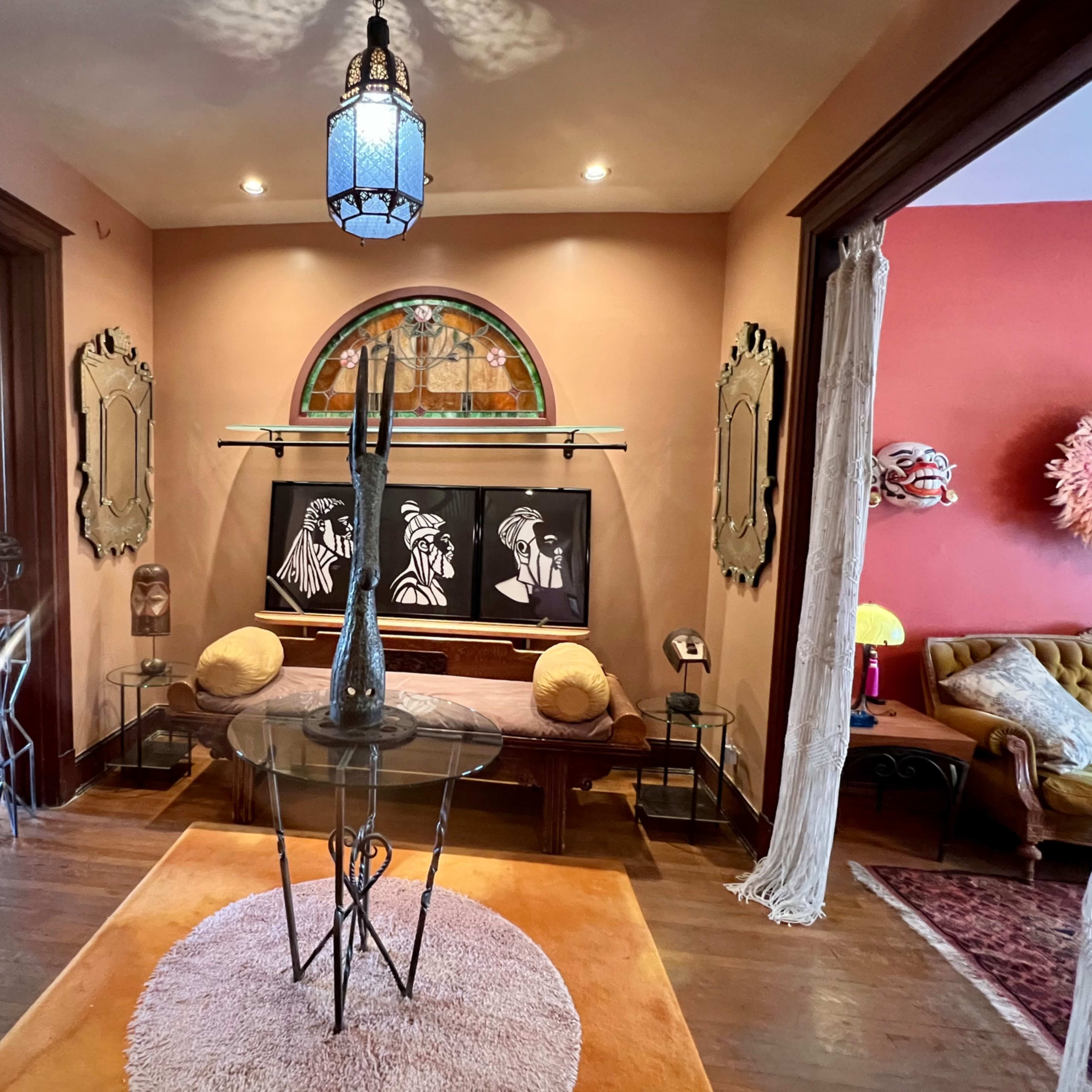The scene depicts a stylish interior featuring a glass table with a tall vase, framed artwork on the wall, and various decorative elements in a warm, inviting room.