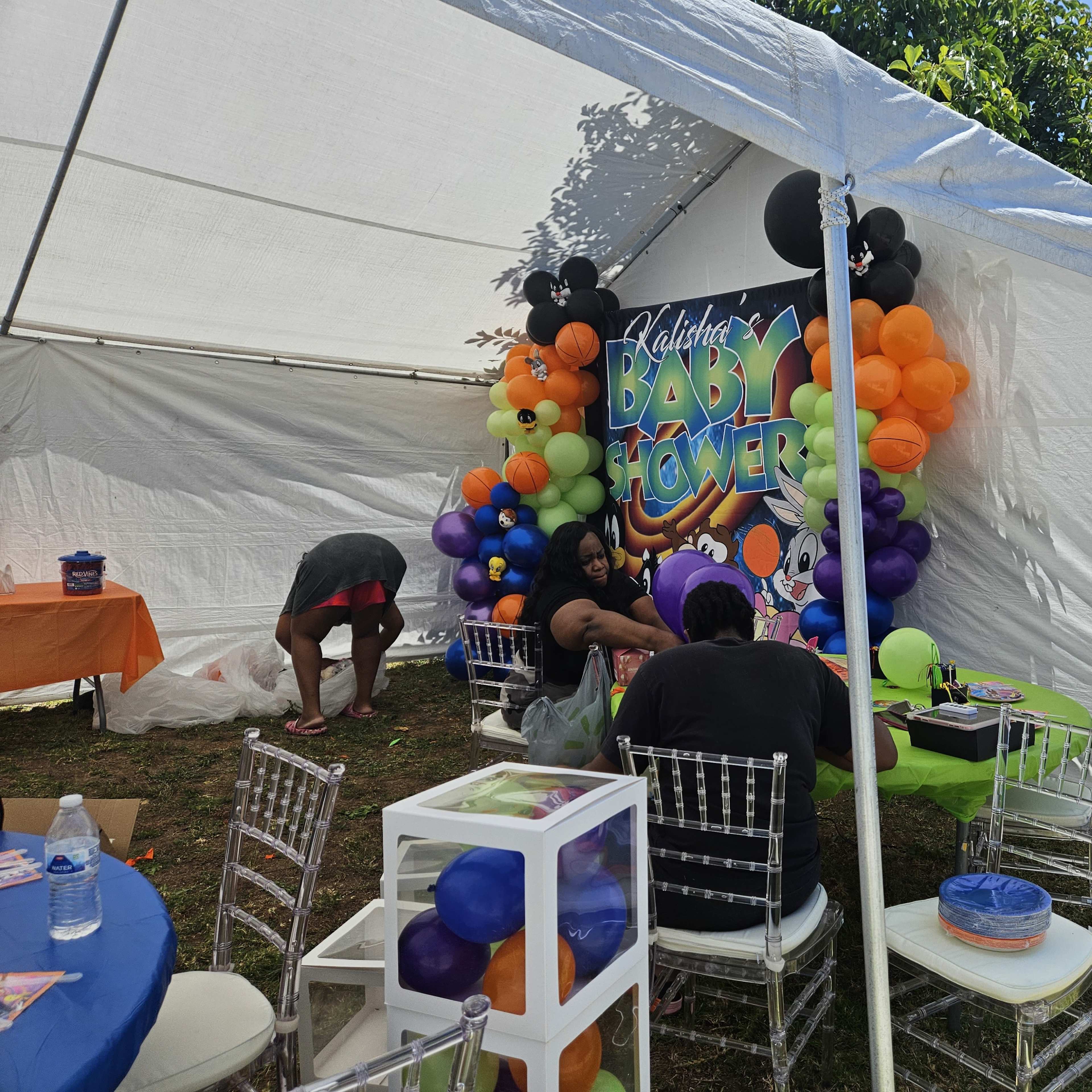 A group is preparing decorations and food under a tent for a baby shower, featuring colorful balloons and a themed backdrop.