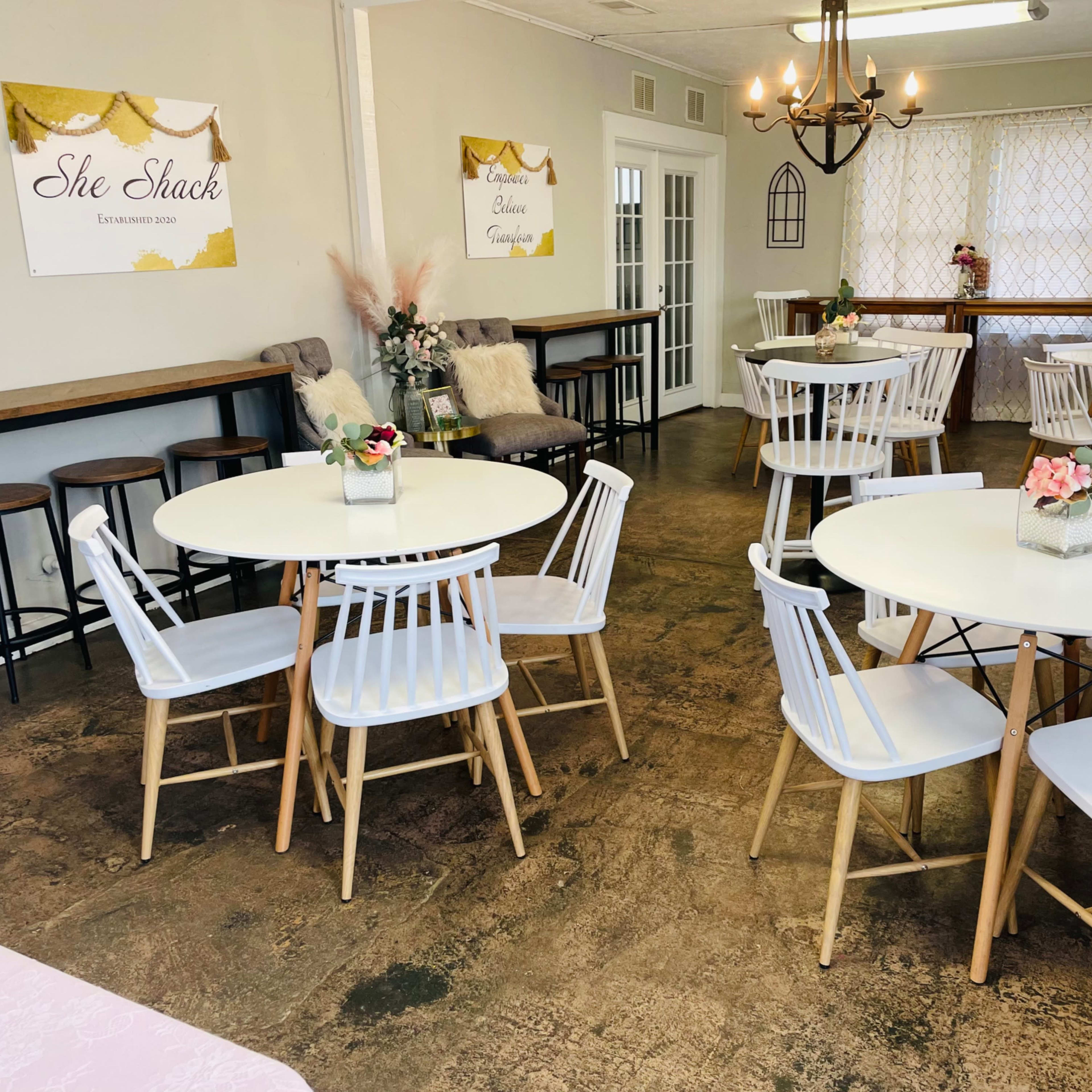 A cozy café interior with white tables and chairs arranged around the space, decorated with flower centerpieces and two posters on the walls.