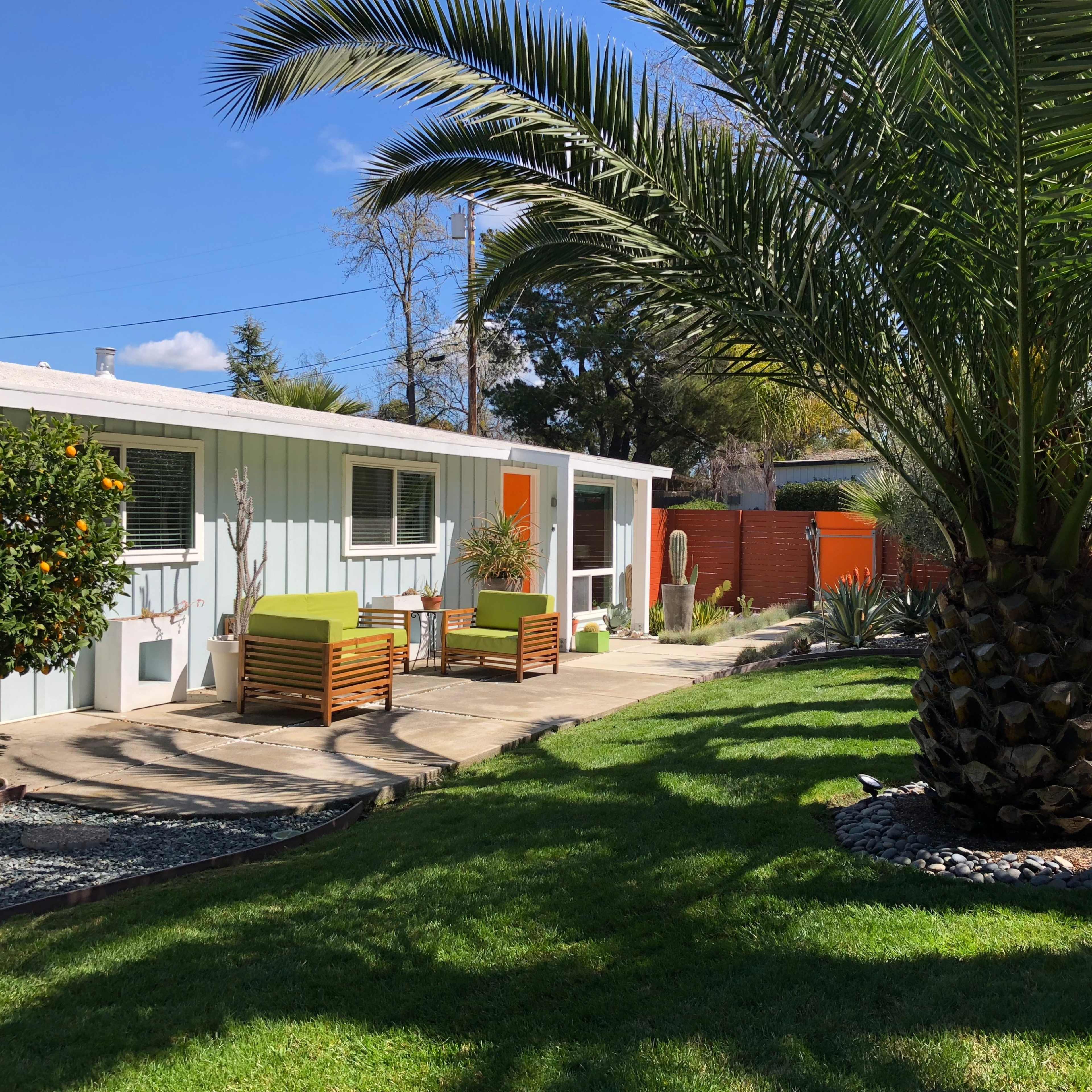 A small house with light blue siding features a patio with two wooden chairs, surrounded by palm trees and landscaped grass.