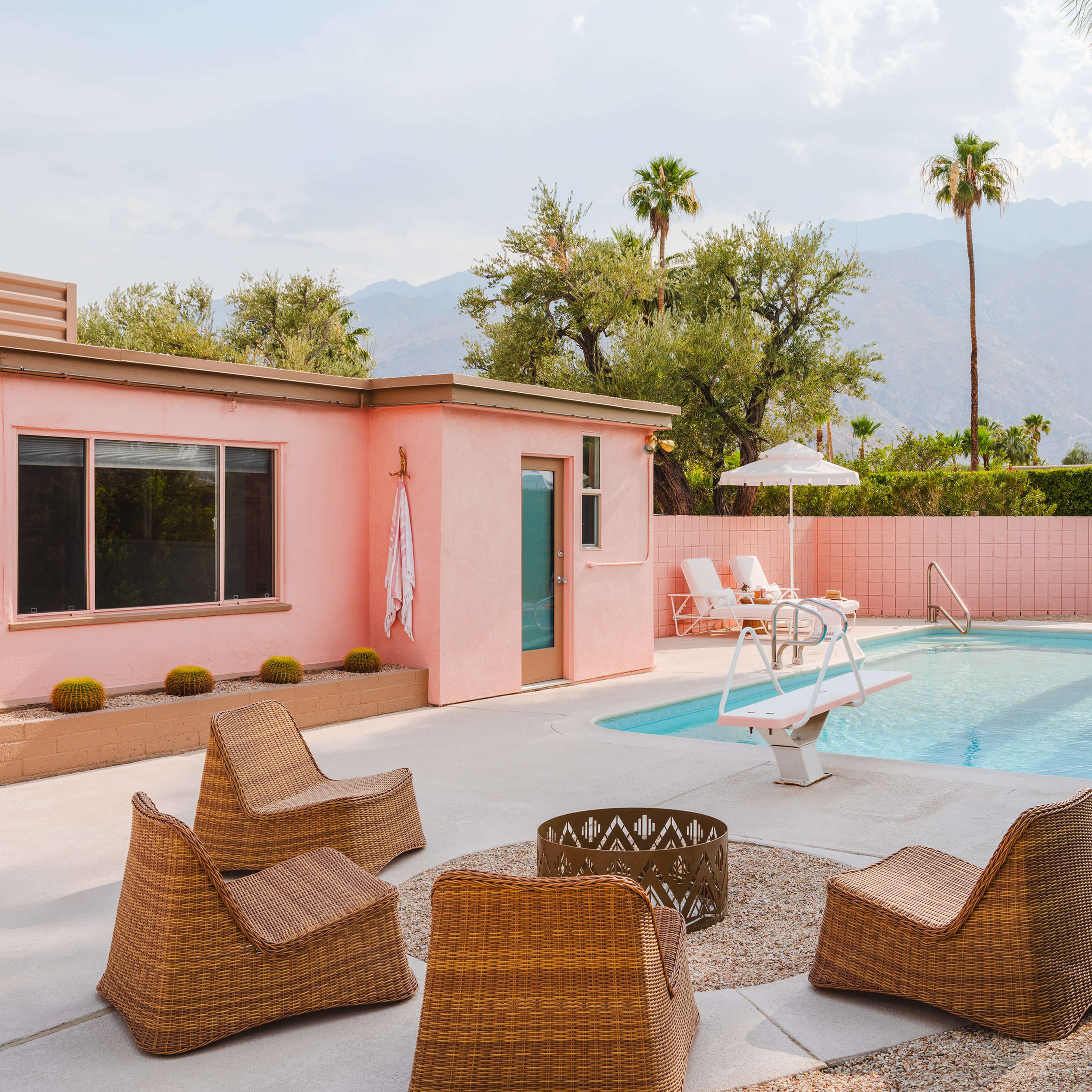 A pink house next to a swimming pool surrounded by lounge chairs and a fire pit, with palm trees and mountains in the background.
