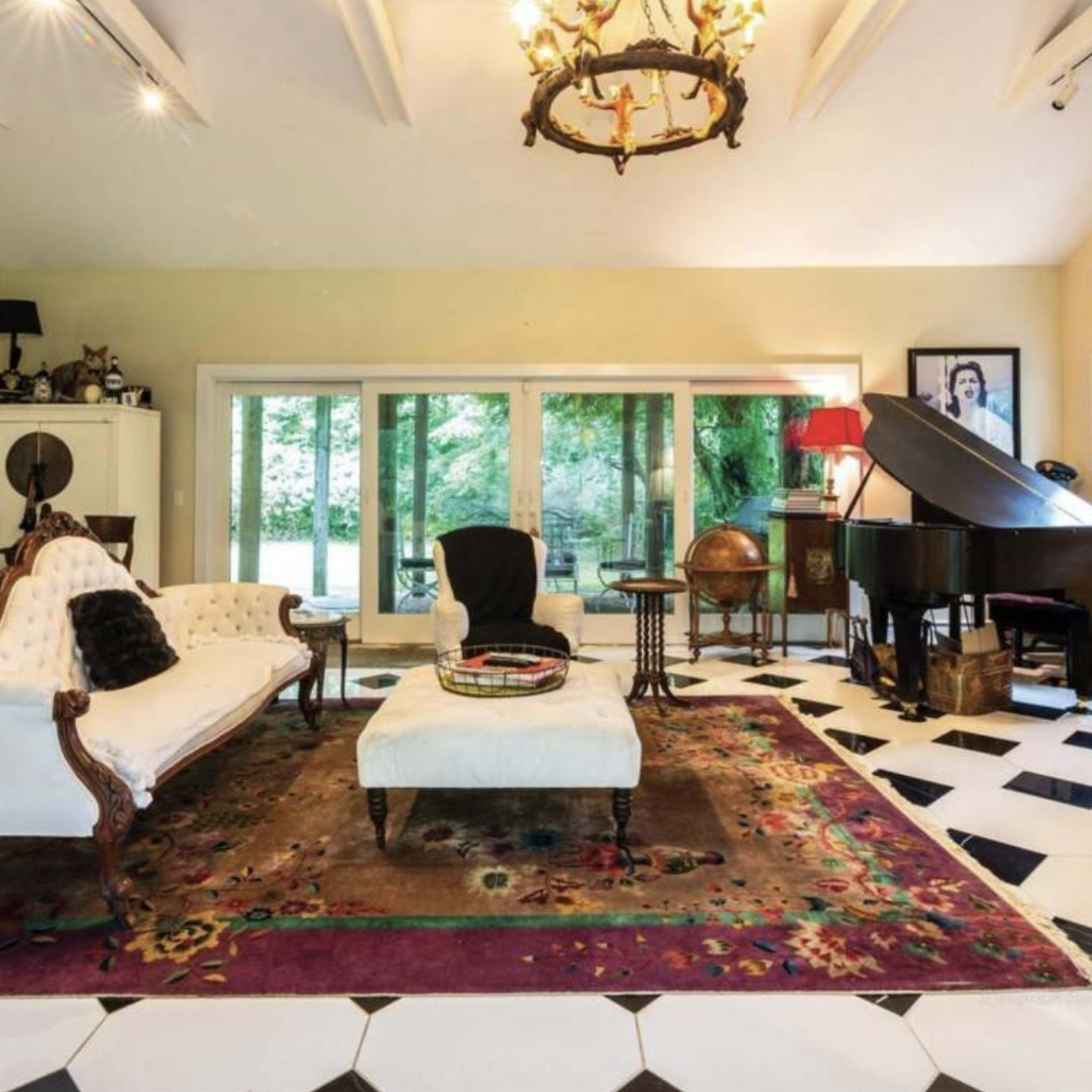 The room features a vintage white sofa, a large area rug, a grand piano, and large windows overlooking greenery, with a checkered black and white floor.