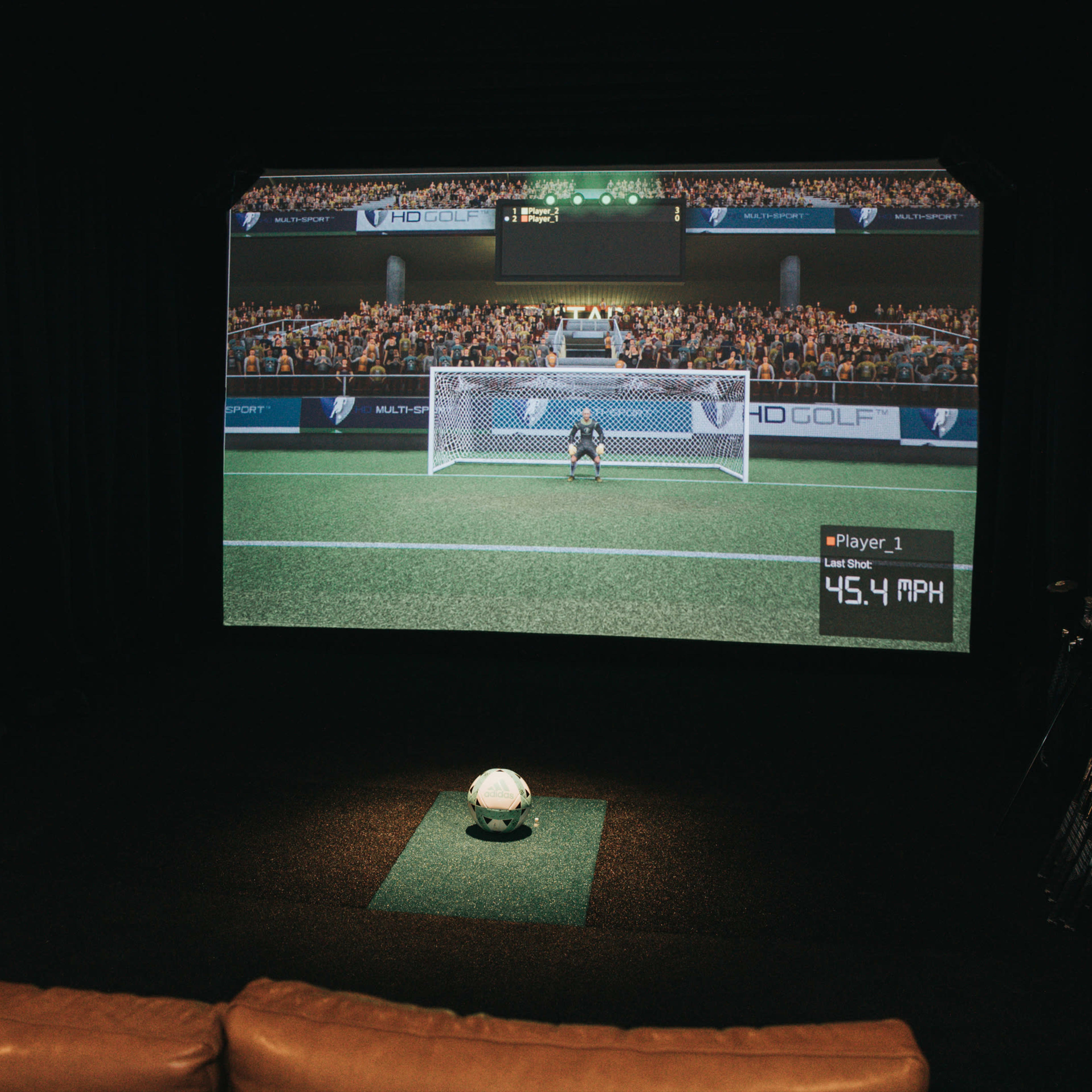 A large projection screen displays a soccer game simulation, with a ball placed on the turf in front of it, while a scoreboard lists player results on the side.