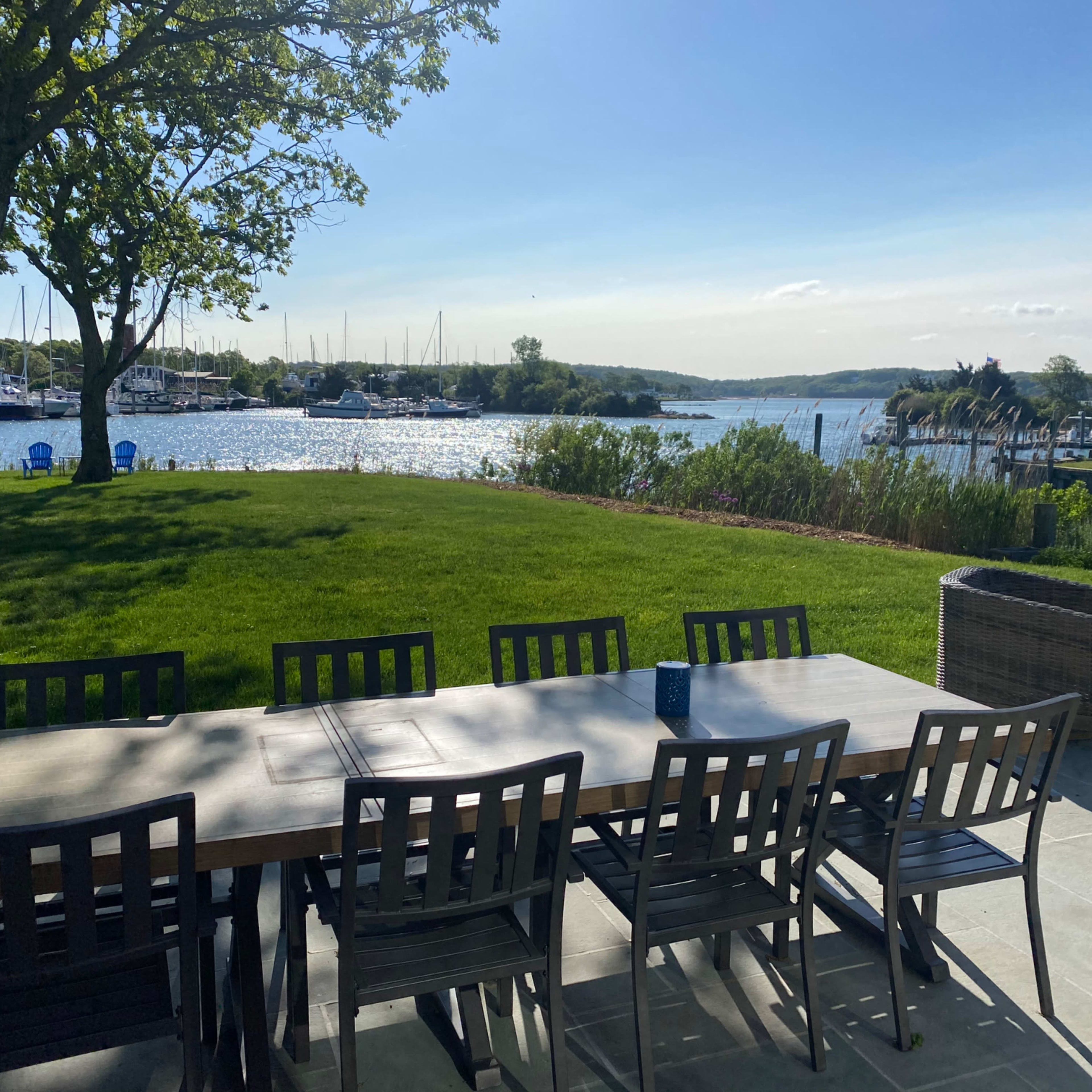 A long table with chairs overlooks a tranquil waterway dotted with boats under a clear sky.