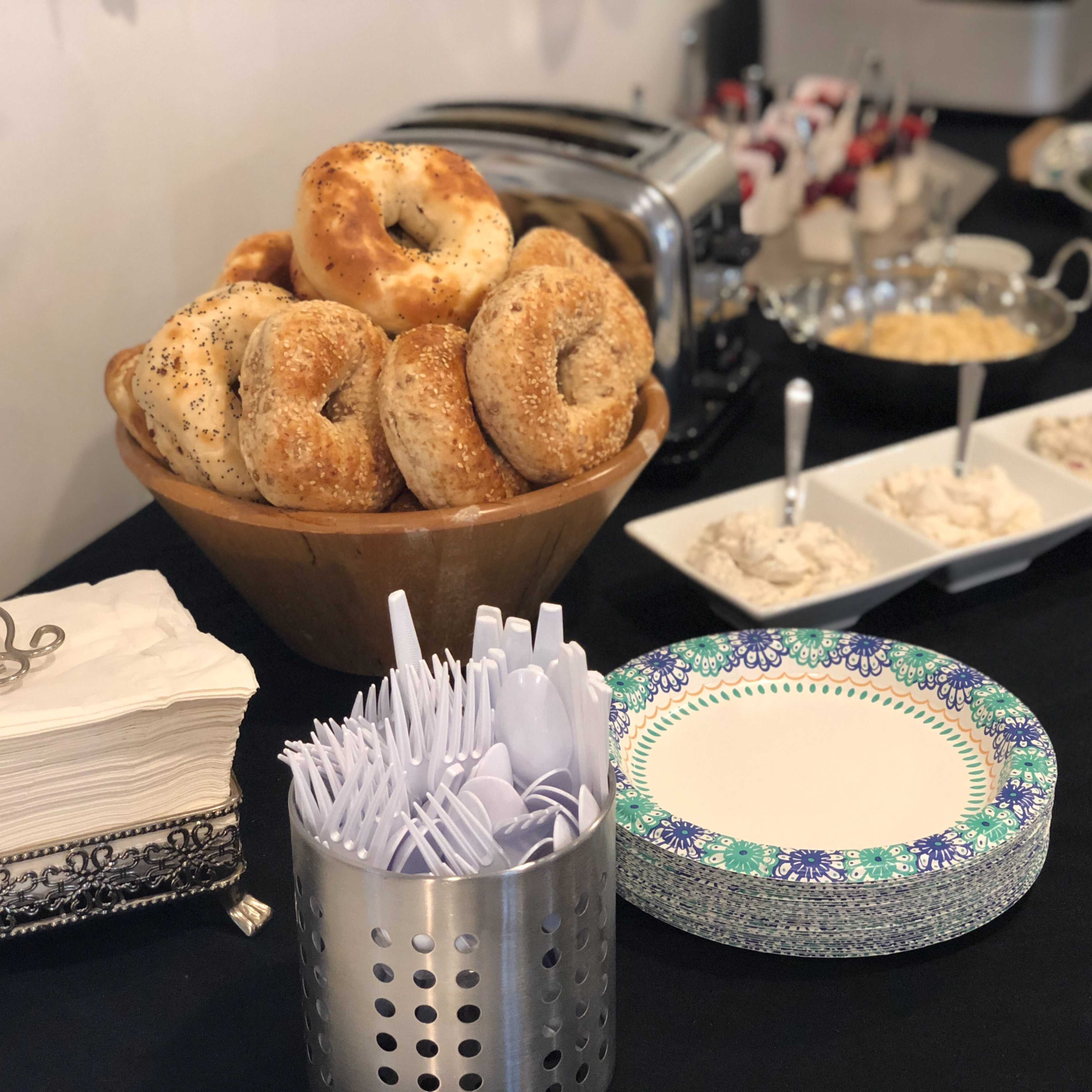 A bowl of bagels is placed next to bowls of cream cheese and a salad bar, accompanied by utensils and plates on a black tablecloth.