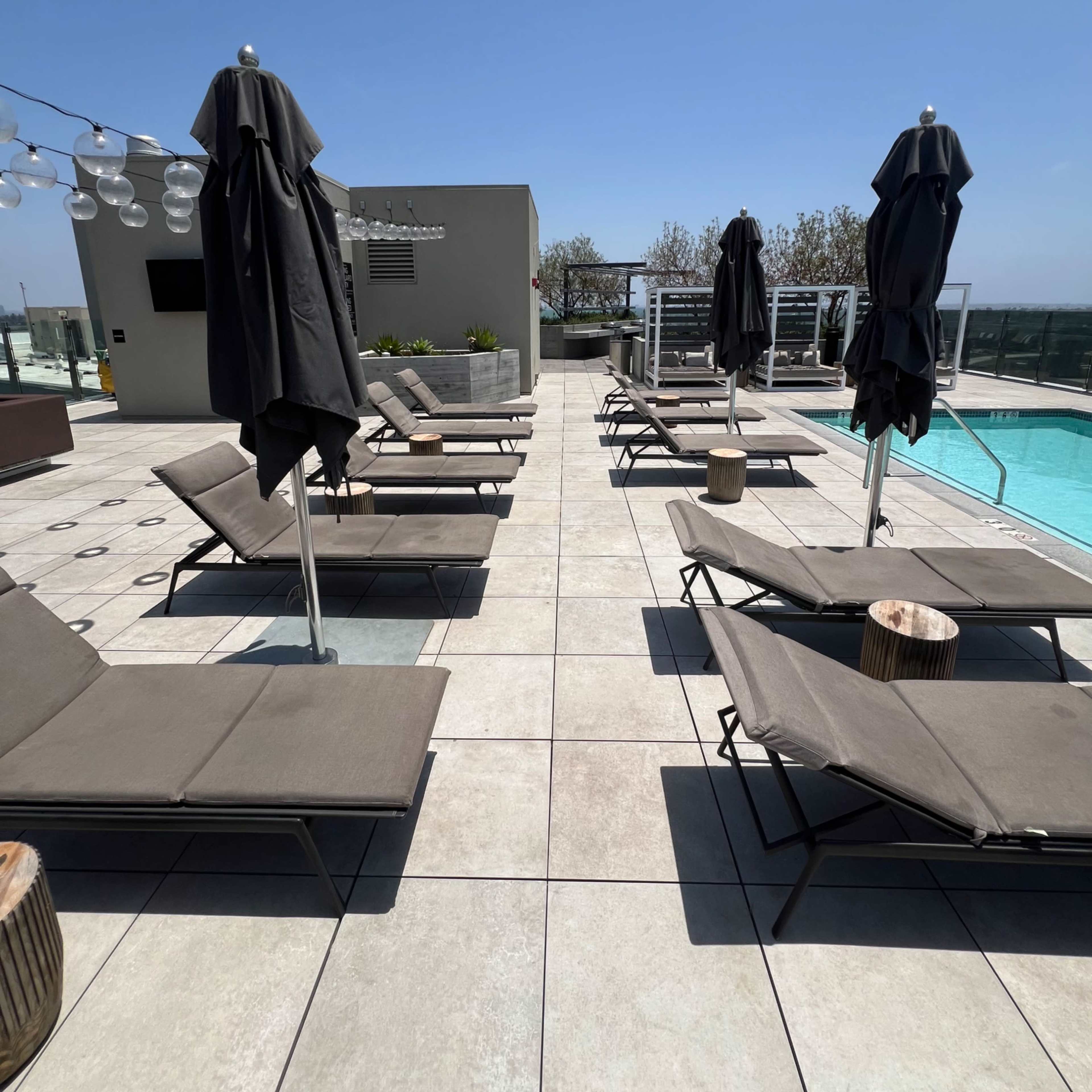 A rooftop pool area features several lounge chairs with umbrellas arranged along the tiled floor beside a swimming pool.