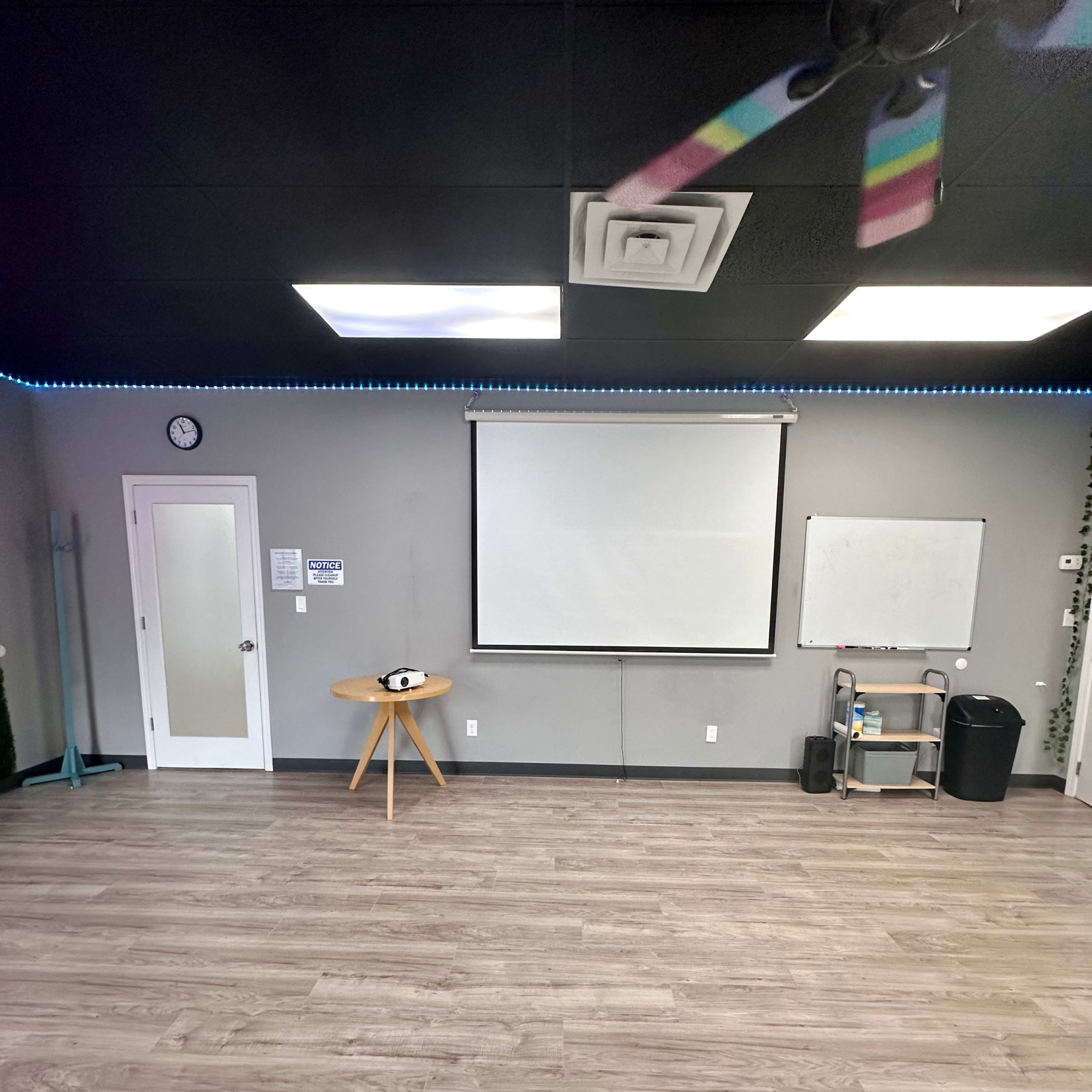 A sparsely decorated room with a large screen, a whiteboard, a clock, and minimal furniture against a gray wall.