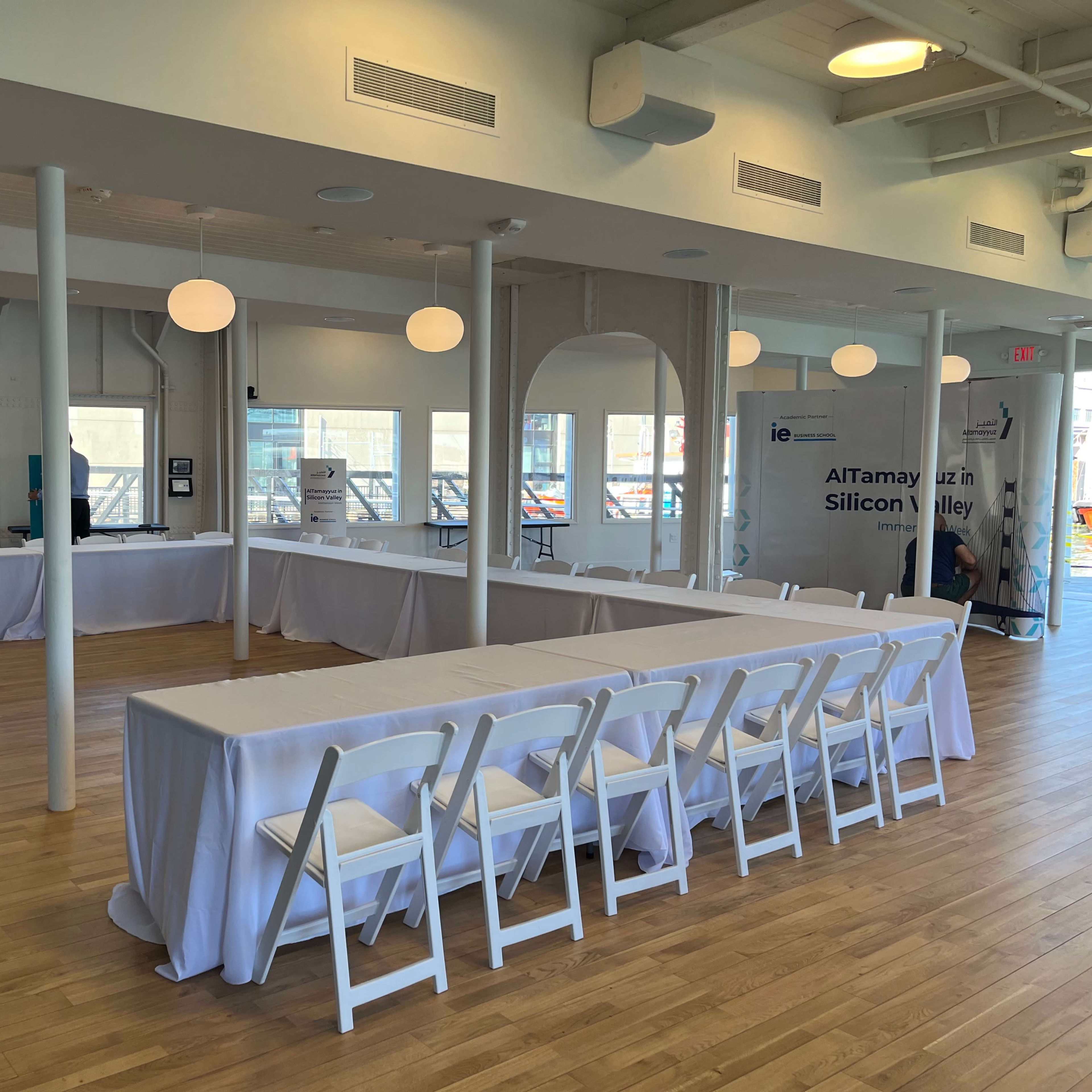 A spacious, well-lit event room with white tables covered in linens and a few promotional banners positioned along the walls.