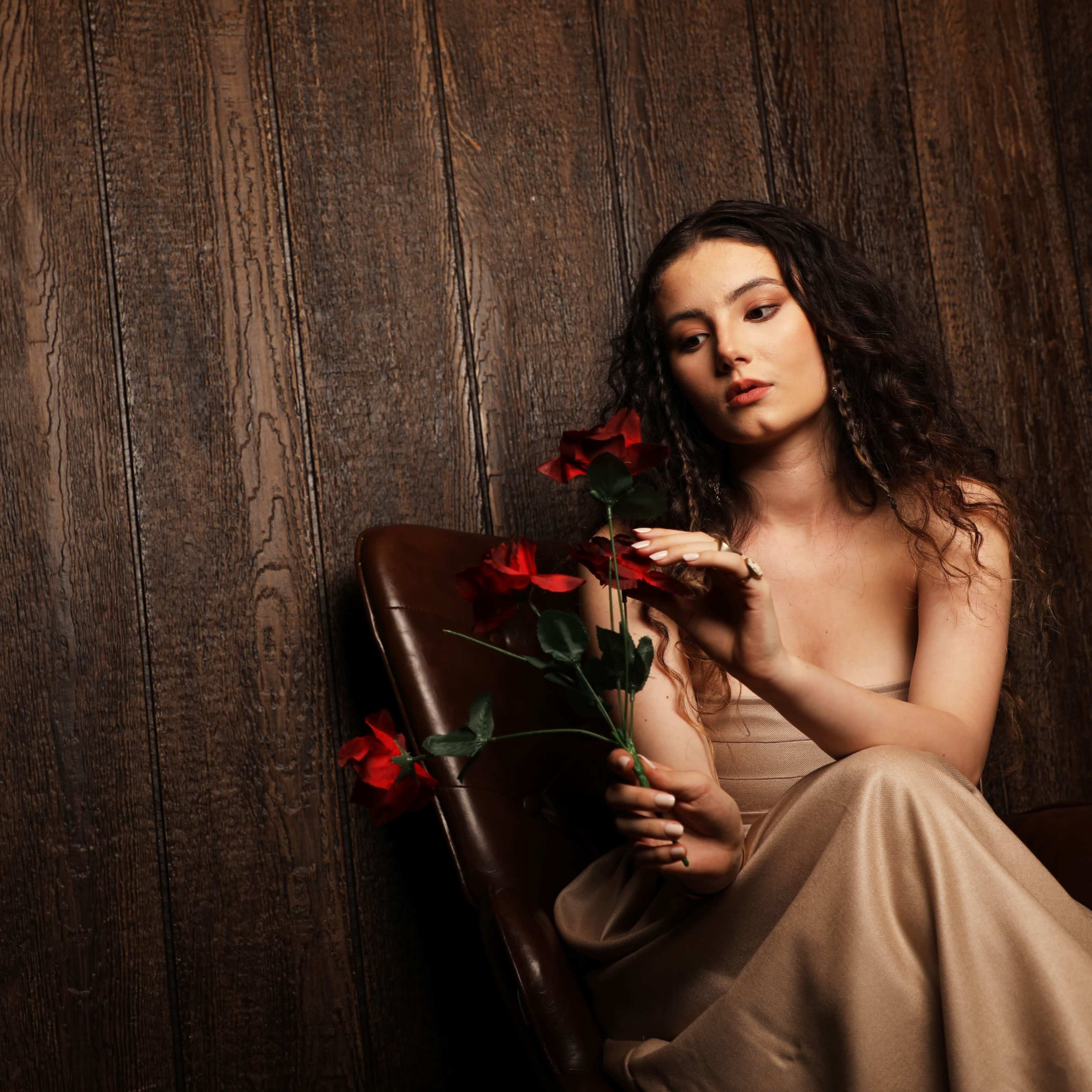A woman in a strapless dress sits in a brown chair, holding red roses against a wooden backdrop.