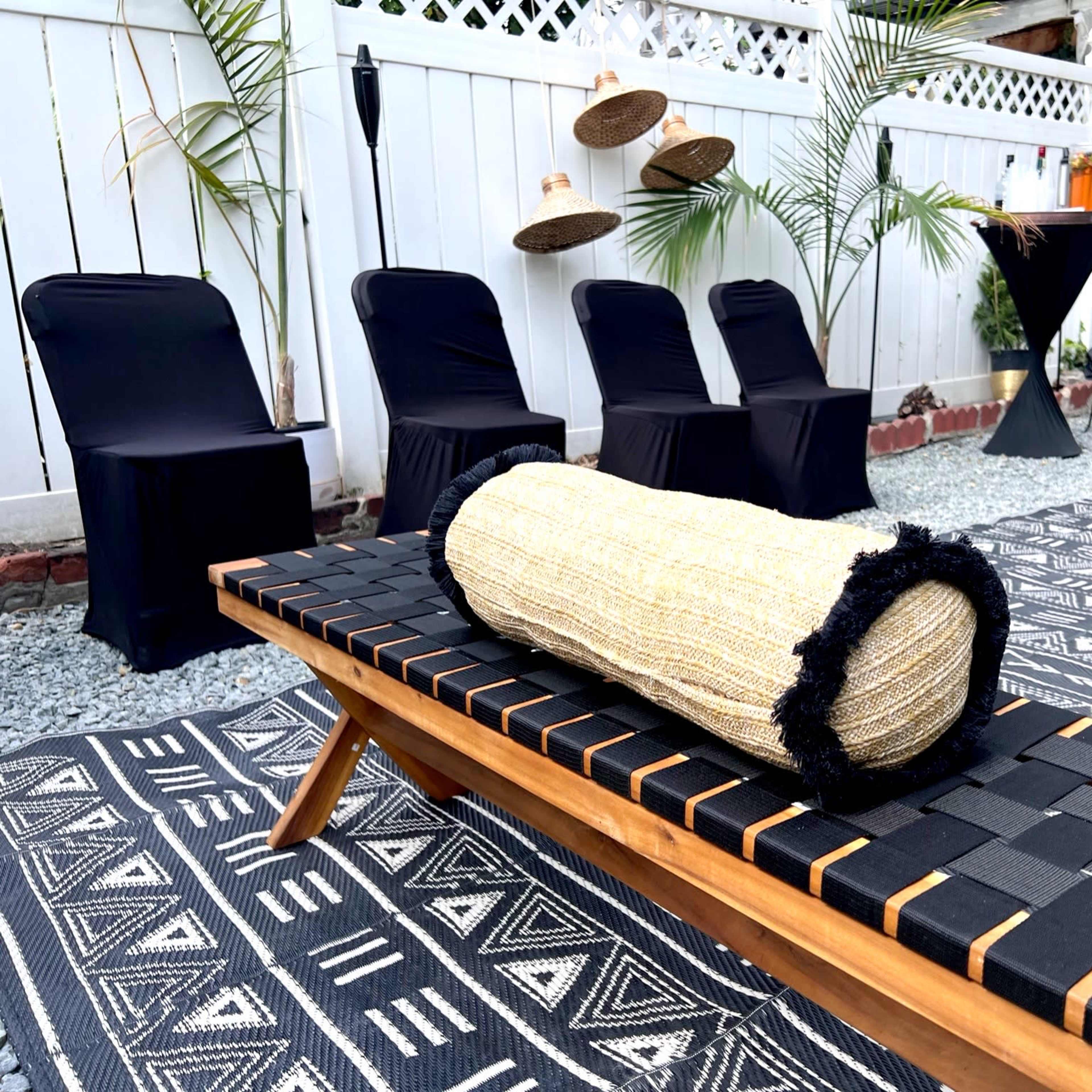 A wooden lounge chair with a cylindrical woven cushion is placed on a patterned outdoor rug surrounded by black chairs and decorative items.