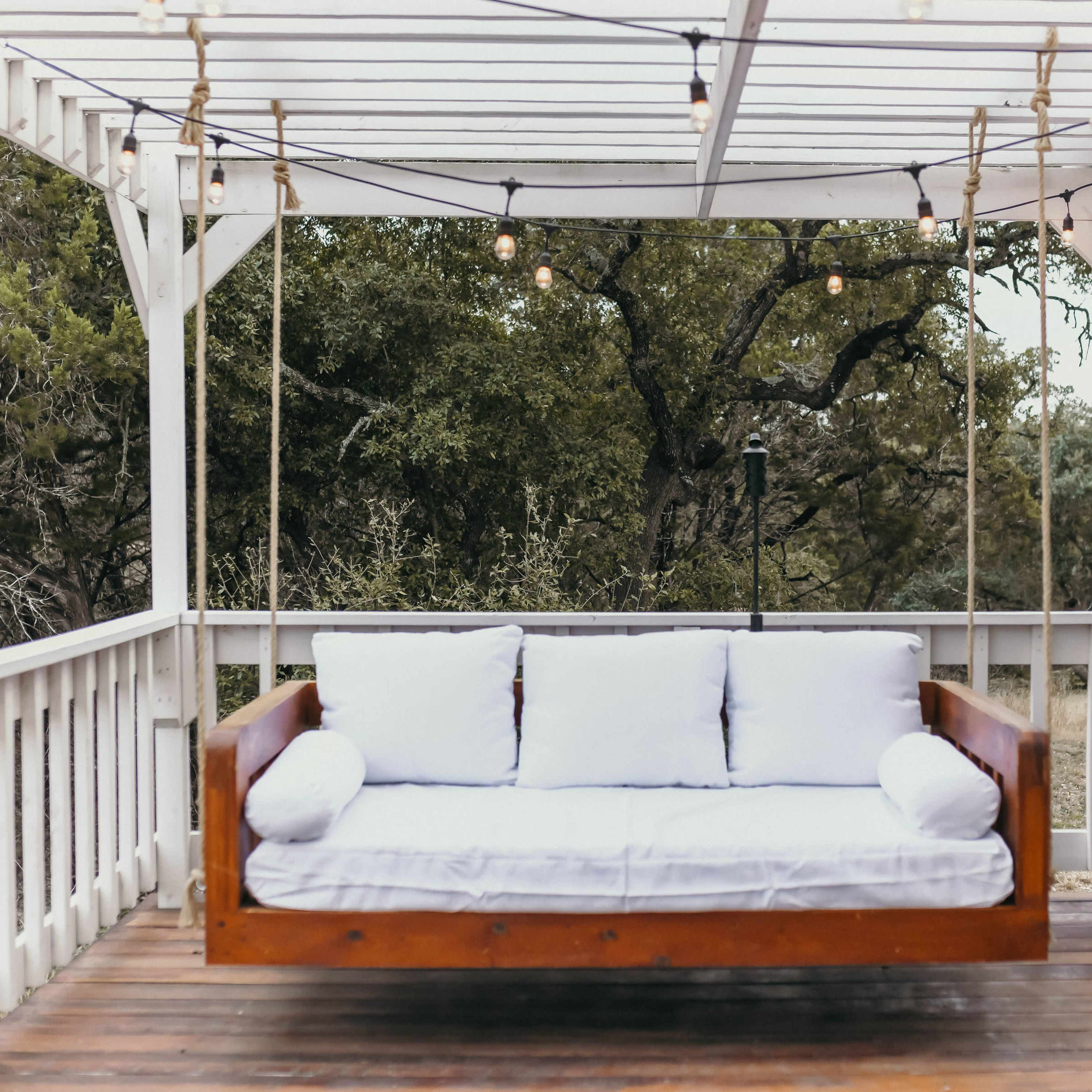 A wooden swing bed with white cushions hangs under a covered patio surrounded by trees.