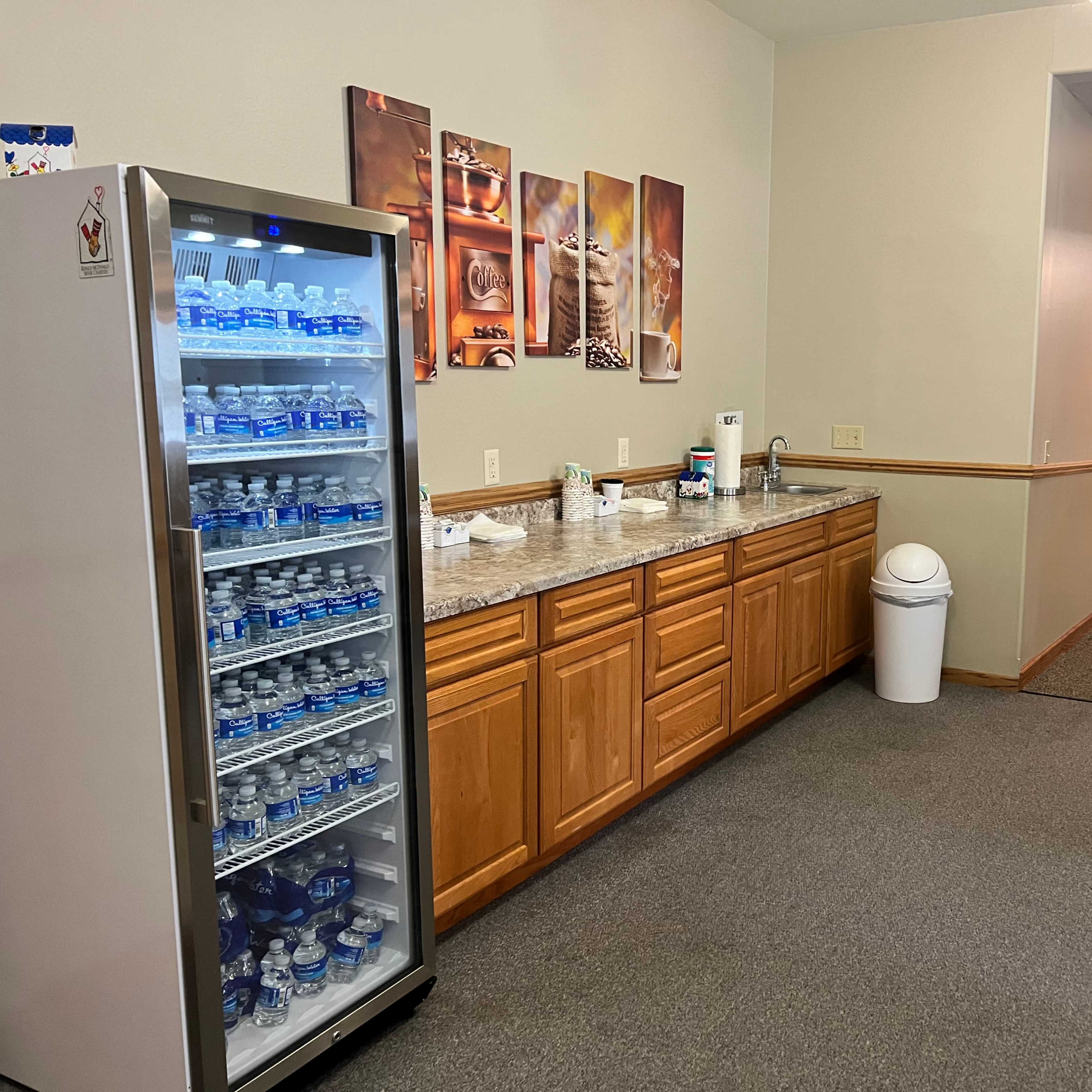A kitchenette area with a refrigerator filled with bottled water, a countertop with coffee supplies, and a hallway leading to another room.