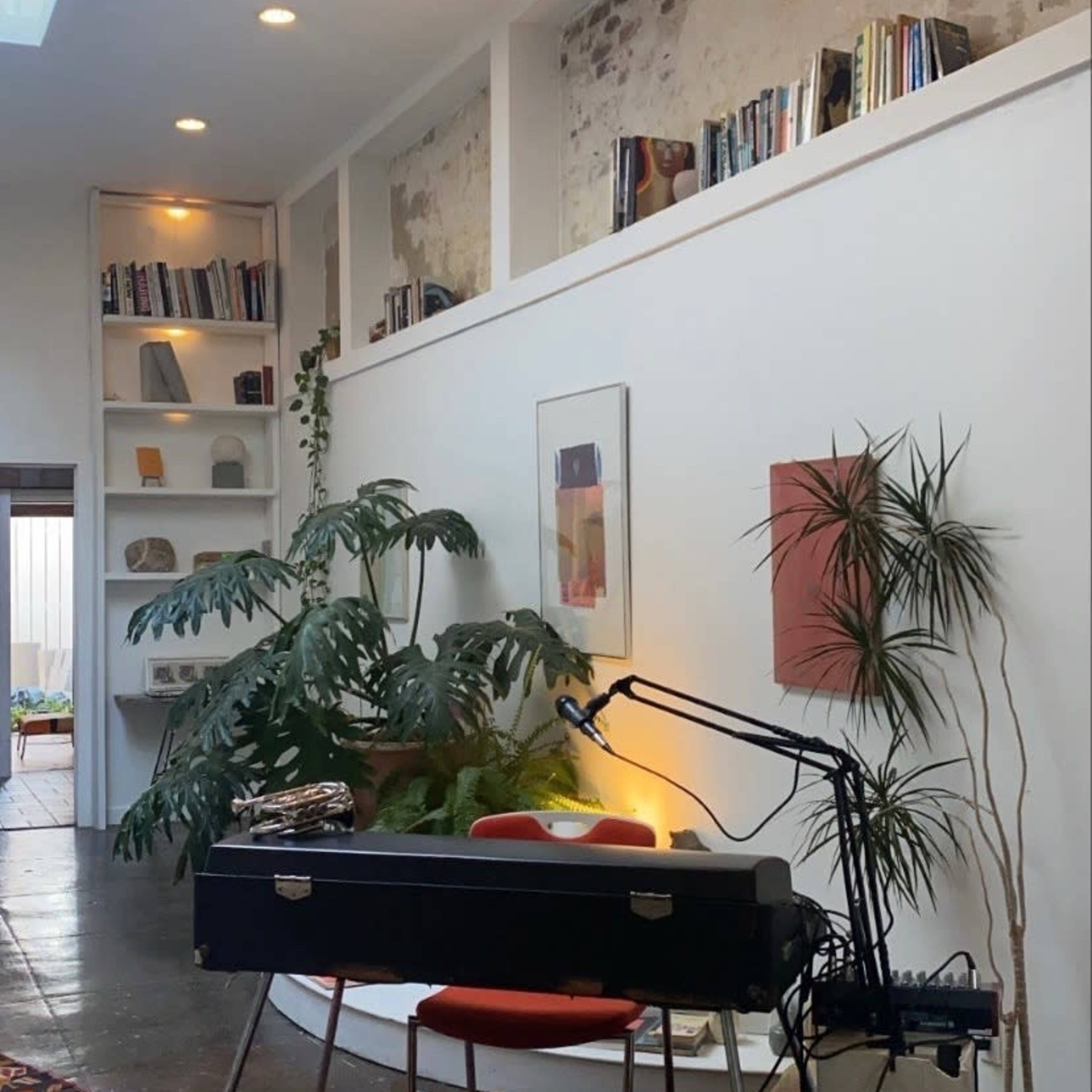 A spacious interior with a piano, a microphone stand, and a wall-mounted bookshelf, complemented by large plants and natural light from a skylight.