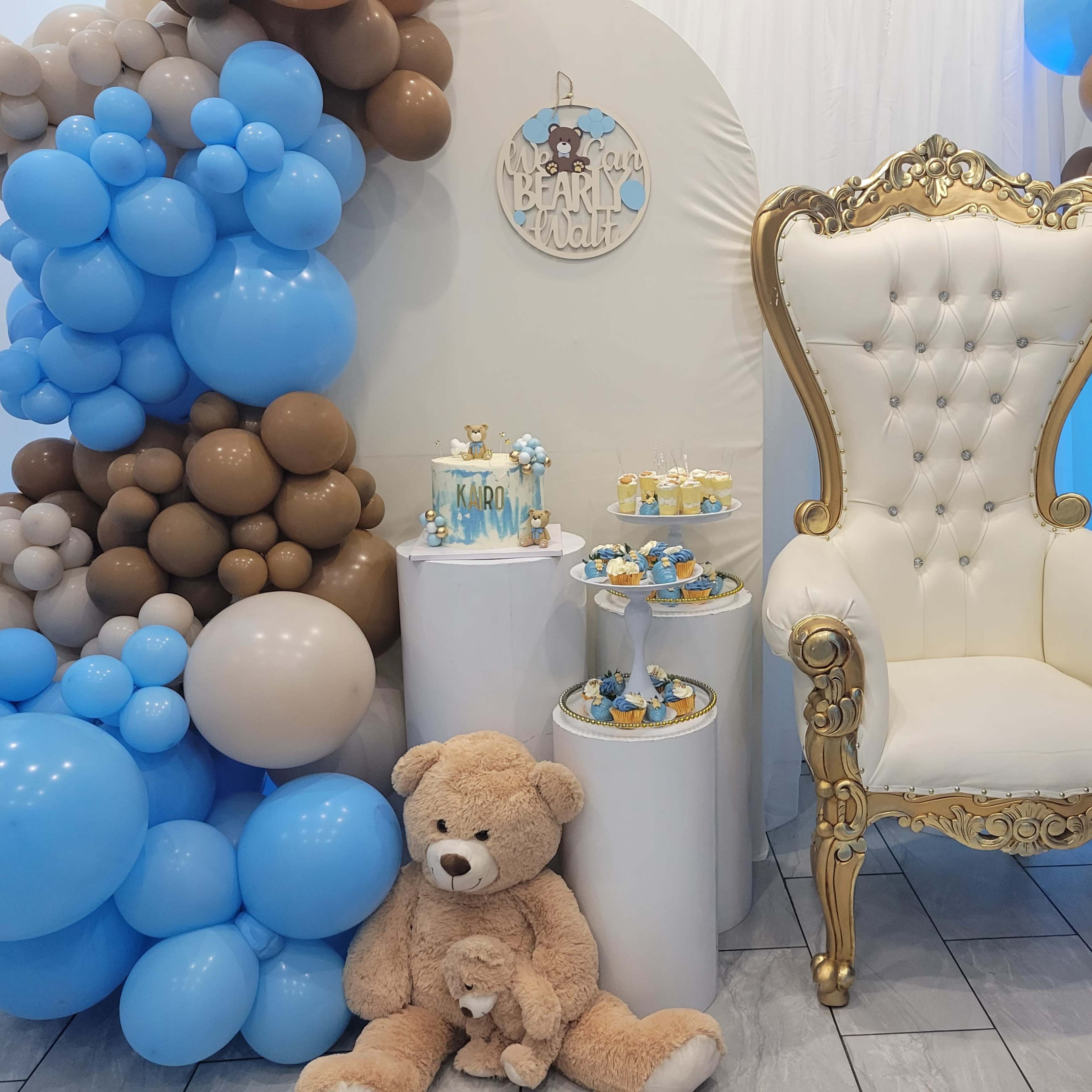 A decorated event space featuring a golden throne chair, a display of dessert treats, and an arrangement of blue and brown balloons.