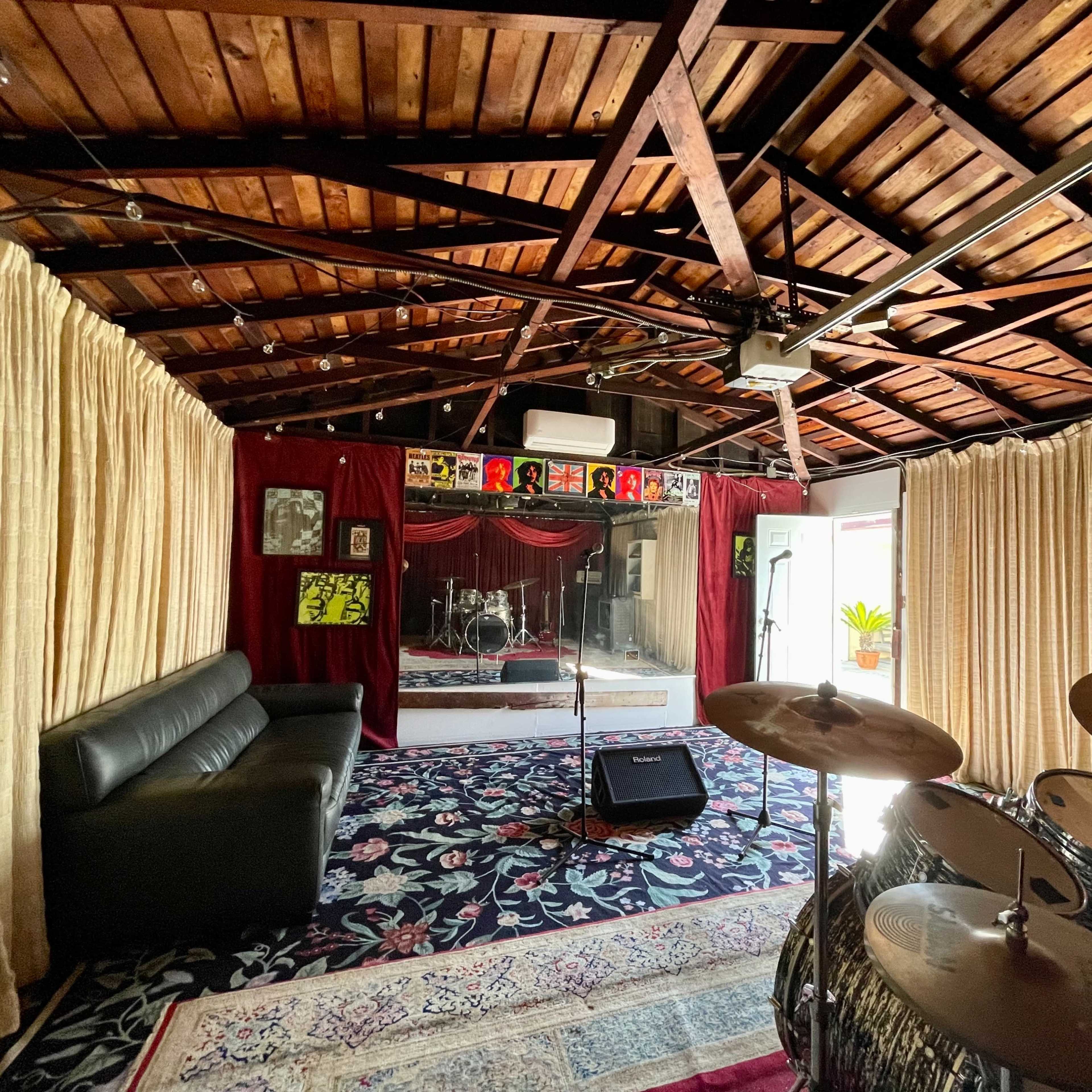 A music rehearsal space with a drum set in the foreground, a sofa, and soundproof curtains, featuring a glassed-in area for a band performance.