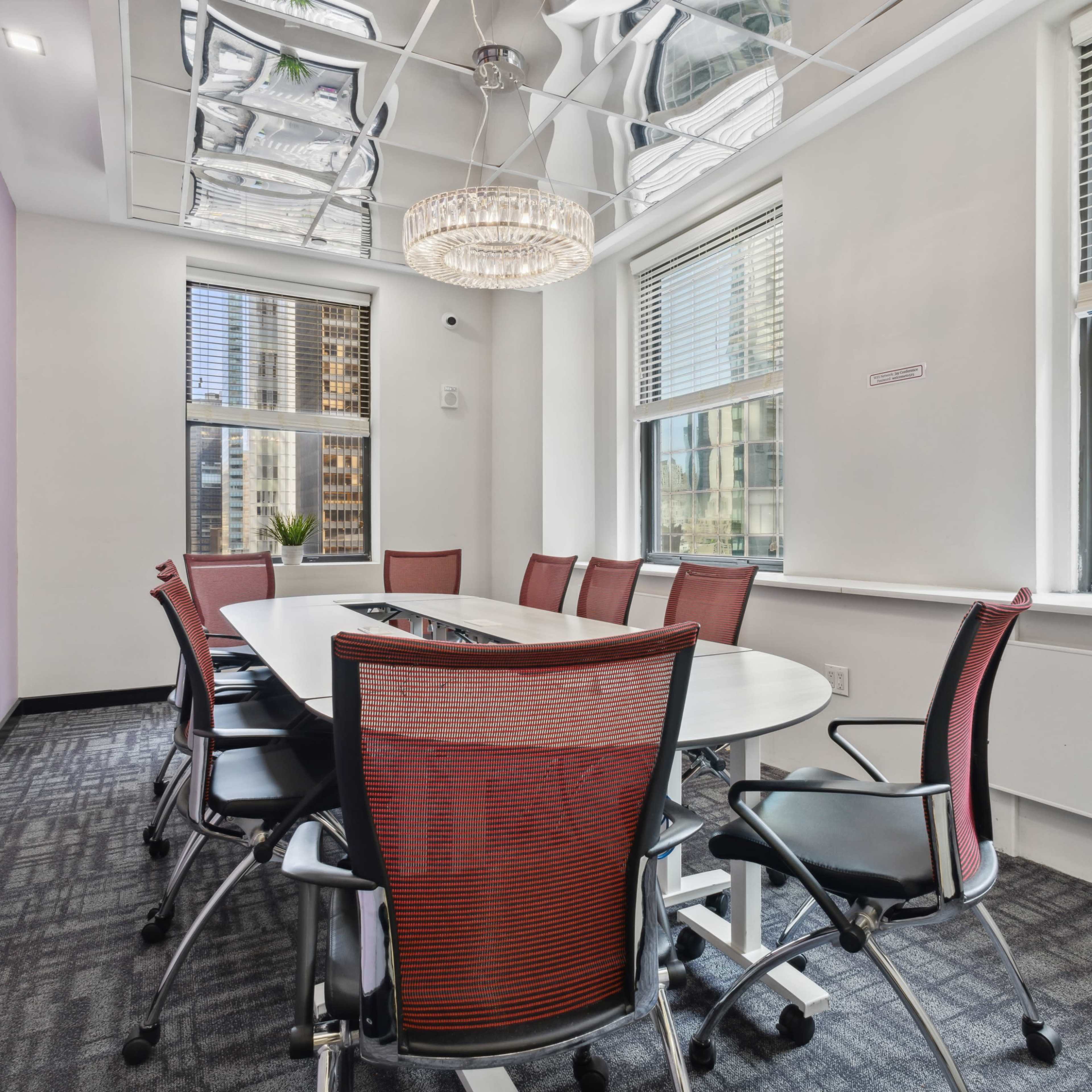 A modern conference room features a large oval table surrounded by red mesh chairs and is illuminated by a chandelier.