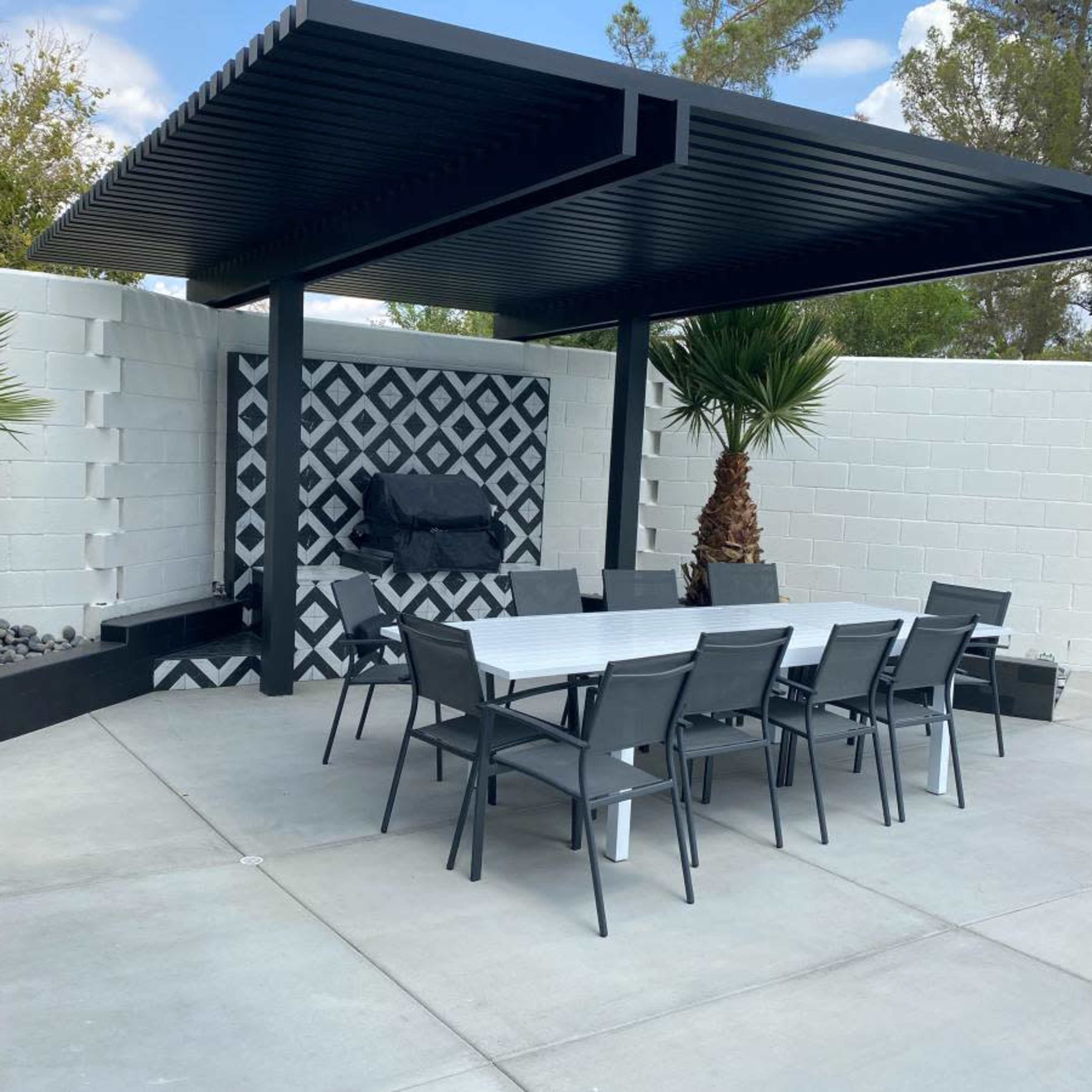 A modern outdoor dining area features a long table and chairs beneath a black pergola, with a patterned accent wall and palm trees nearby.