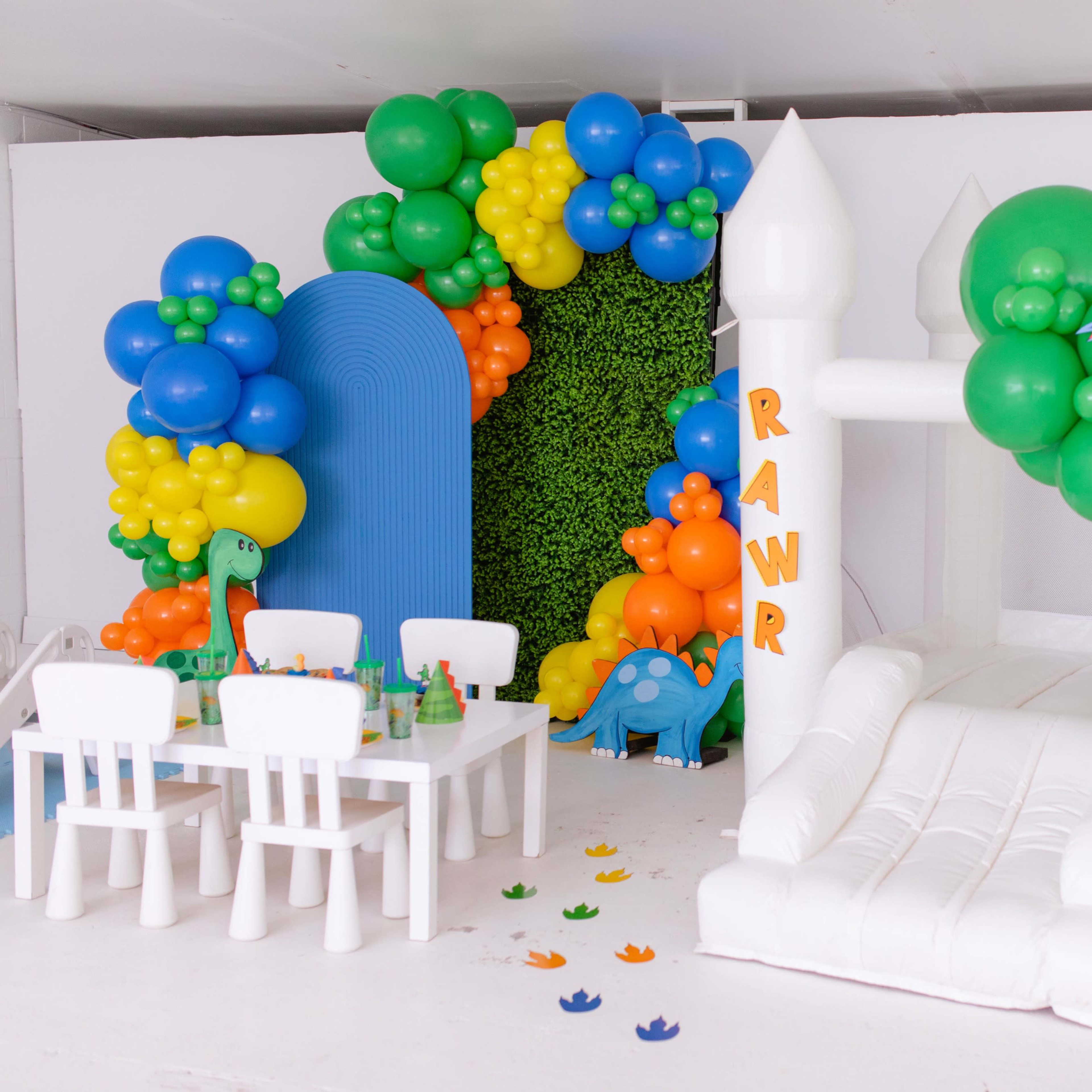 A vibrant party area featuring a white inflatable bounce house, decorated with colorful balloons in a dinosaur theme, alongside a small table set for children.