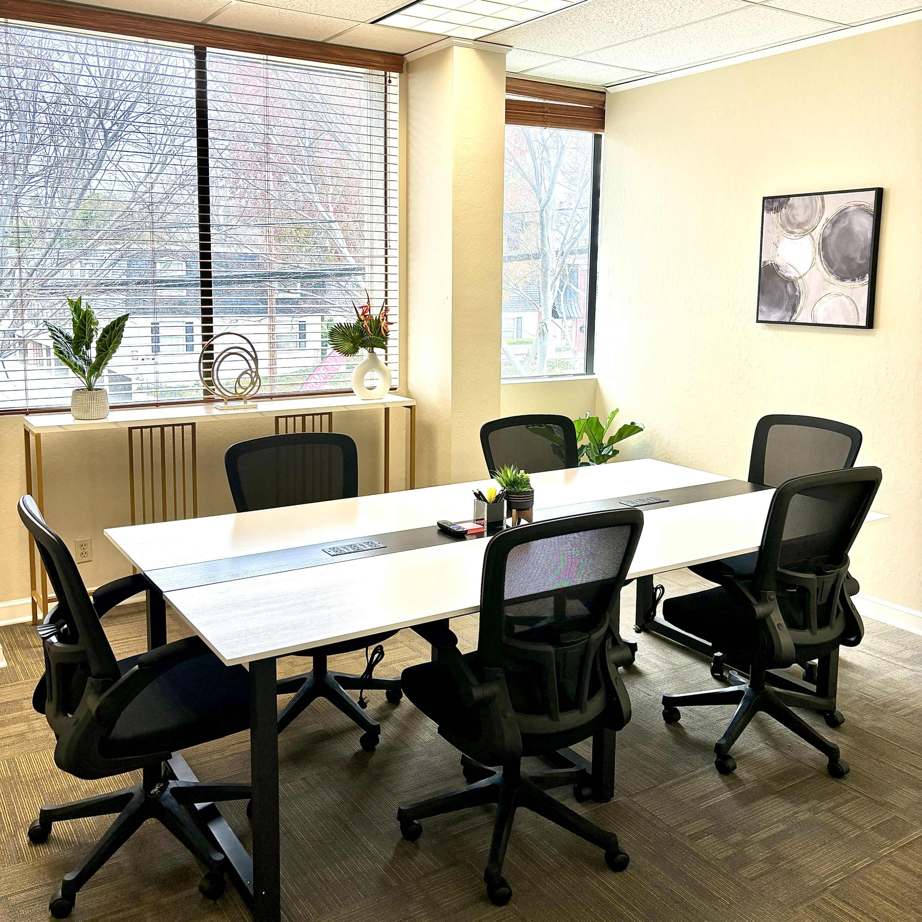 A bright office meeting room features a large white table surrounded by four black chairs, with windows offering a view of outside greenery.