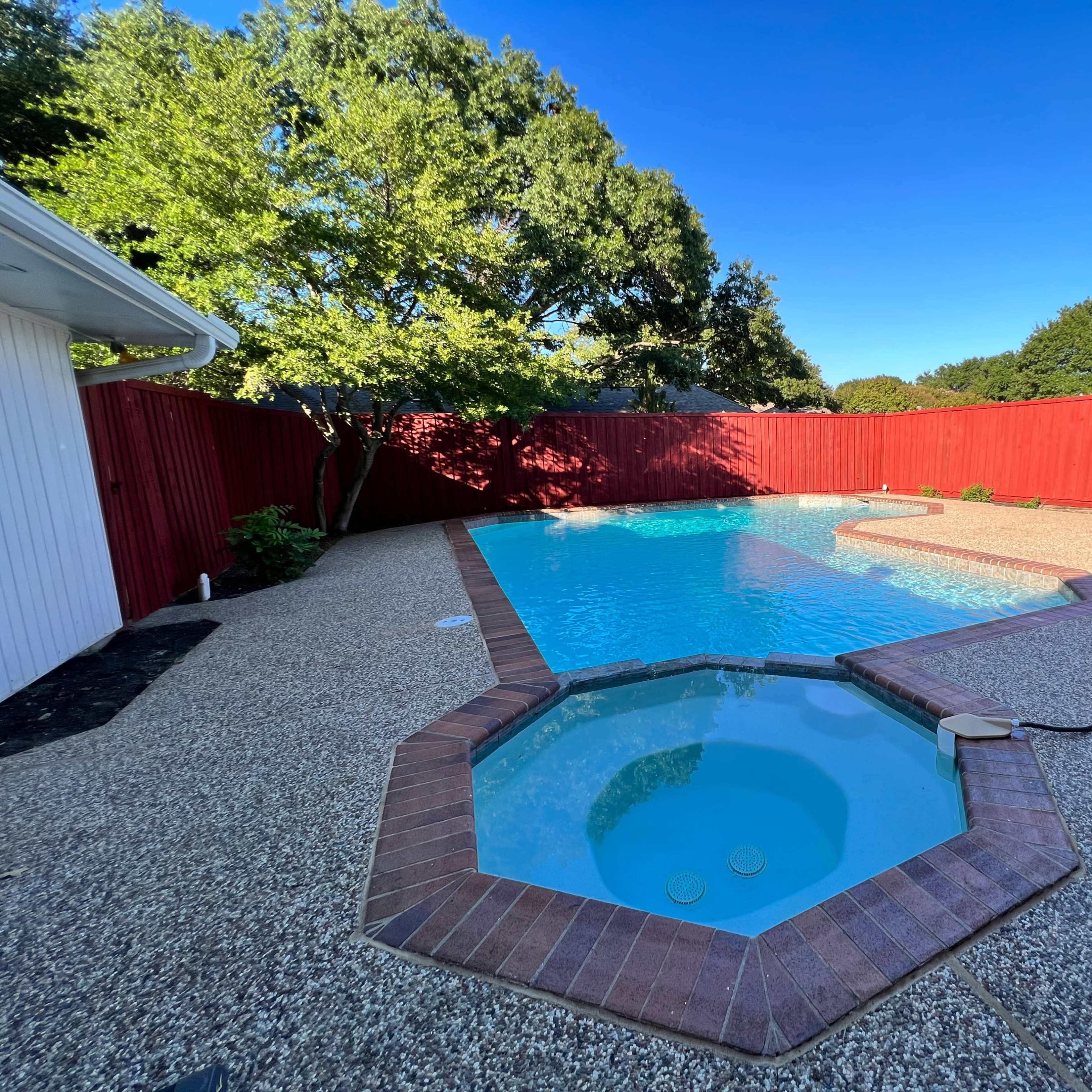A backyard pool area with a shaped pool surrounded by a red fence and patio, featuring a small hot tub on one side.