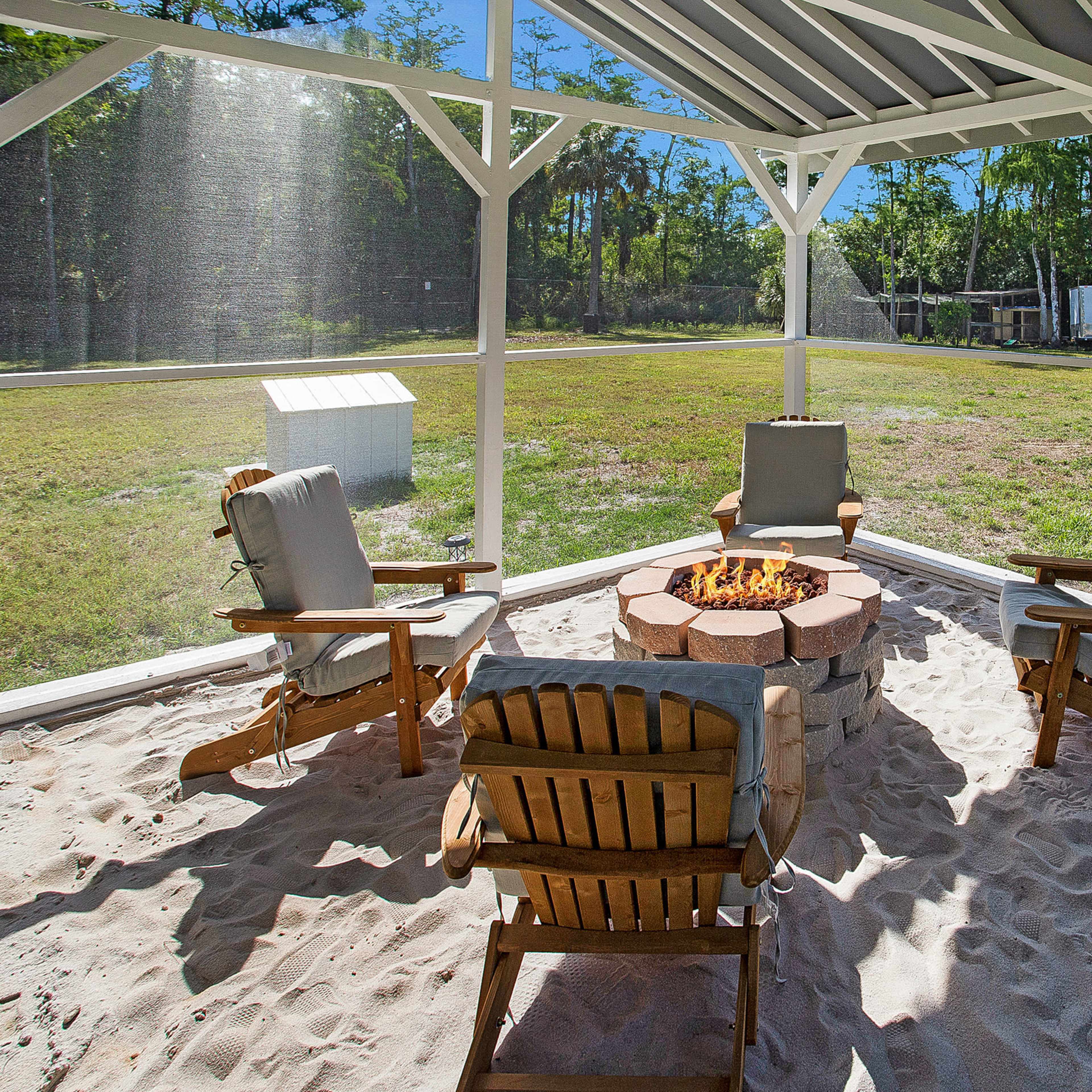 A screened-in outdoor area with a fire pit surrounded by three wooden chairs on sandy ground.