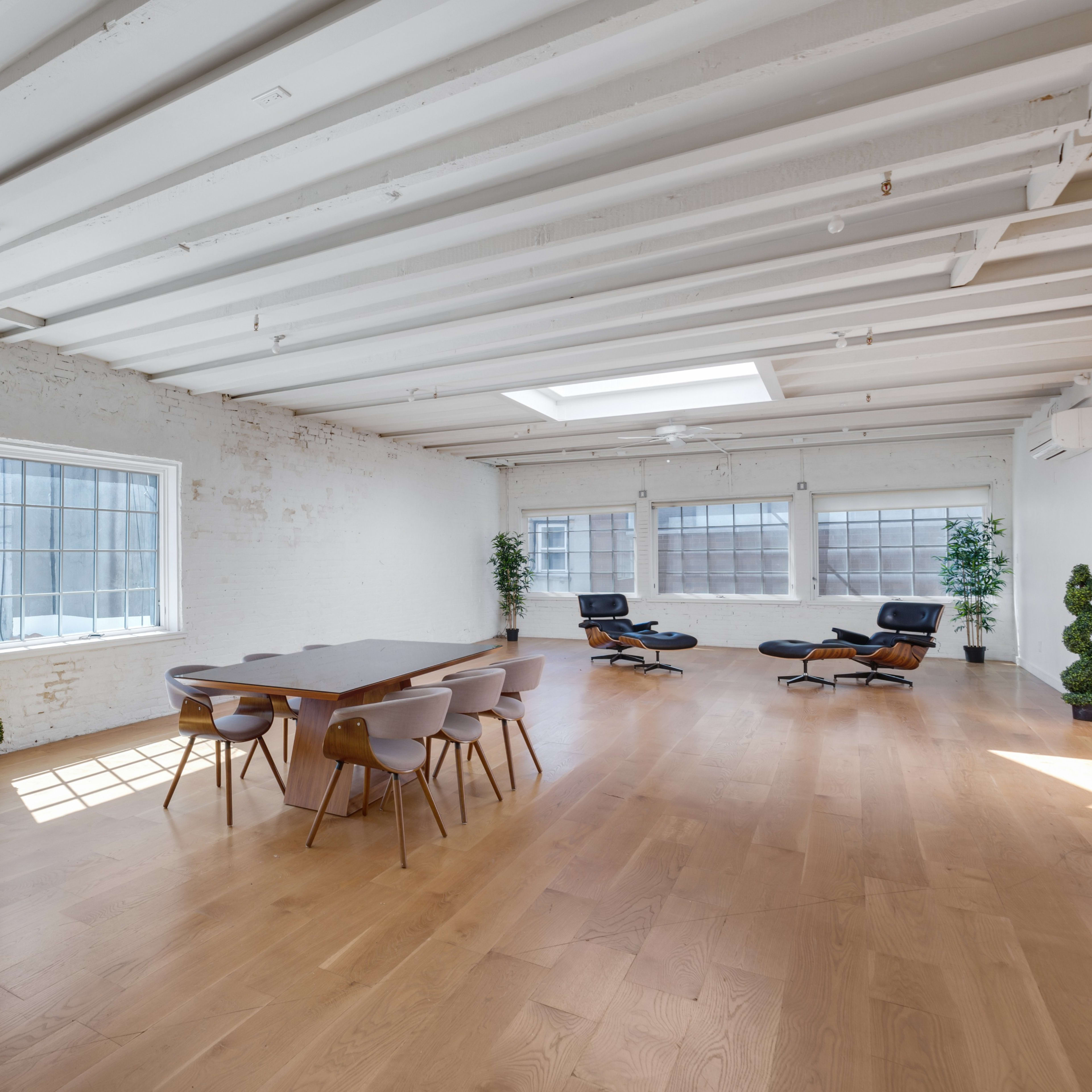 A spacious, brightly lit room features a wooden dining table surrounded by chairs, with large windows and minimalist decor, including potted plants and an office chair in one corner.