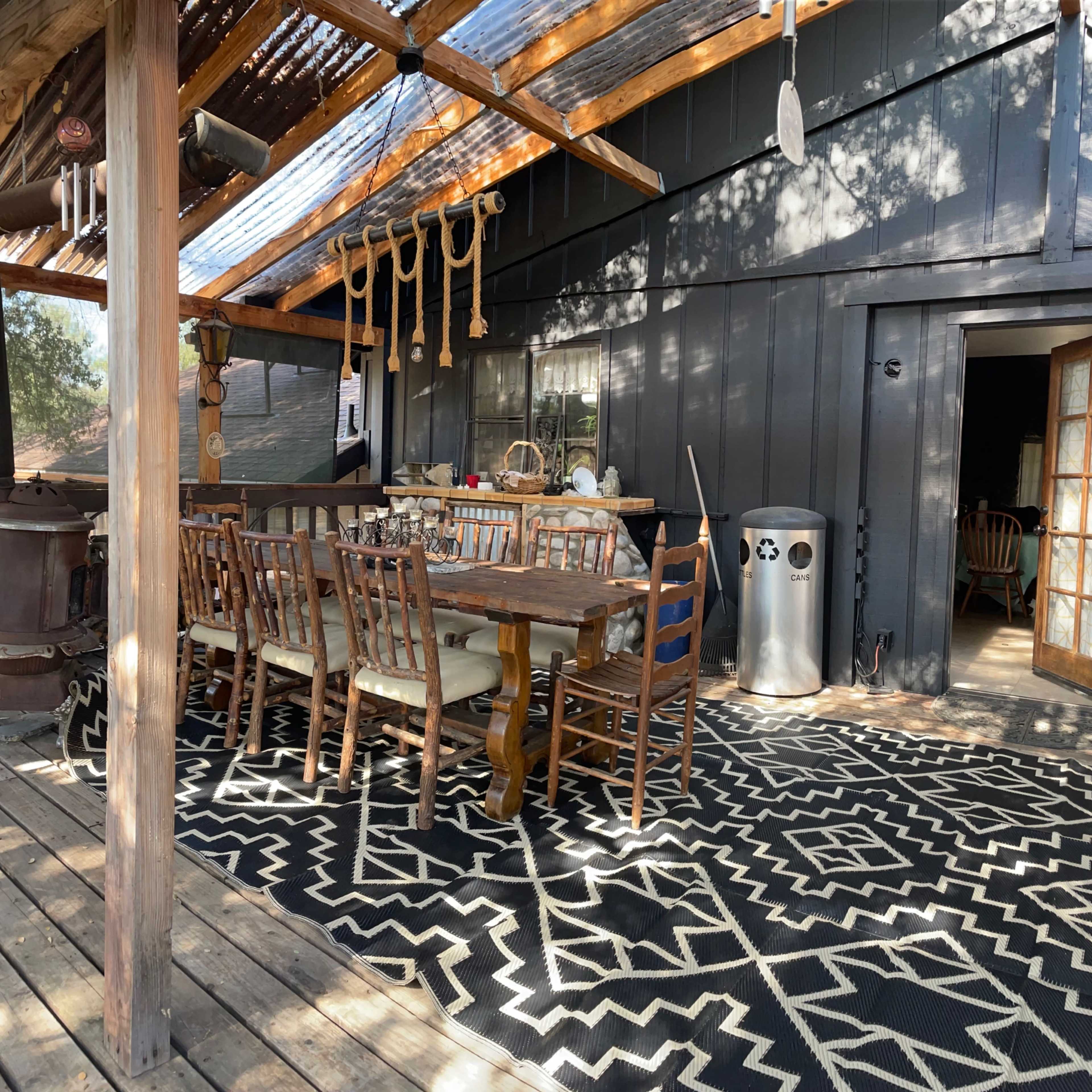 An outdoor dining area with a large wooden table surrounded by chairs, situated under a covered porch with a patterned rug and various decorative items.