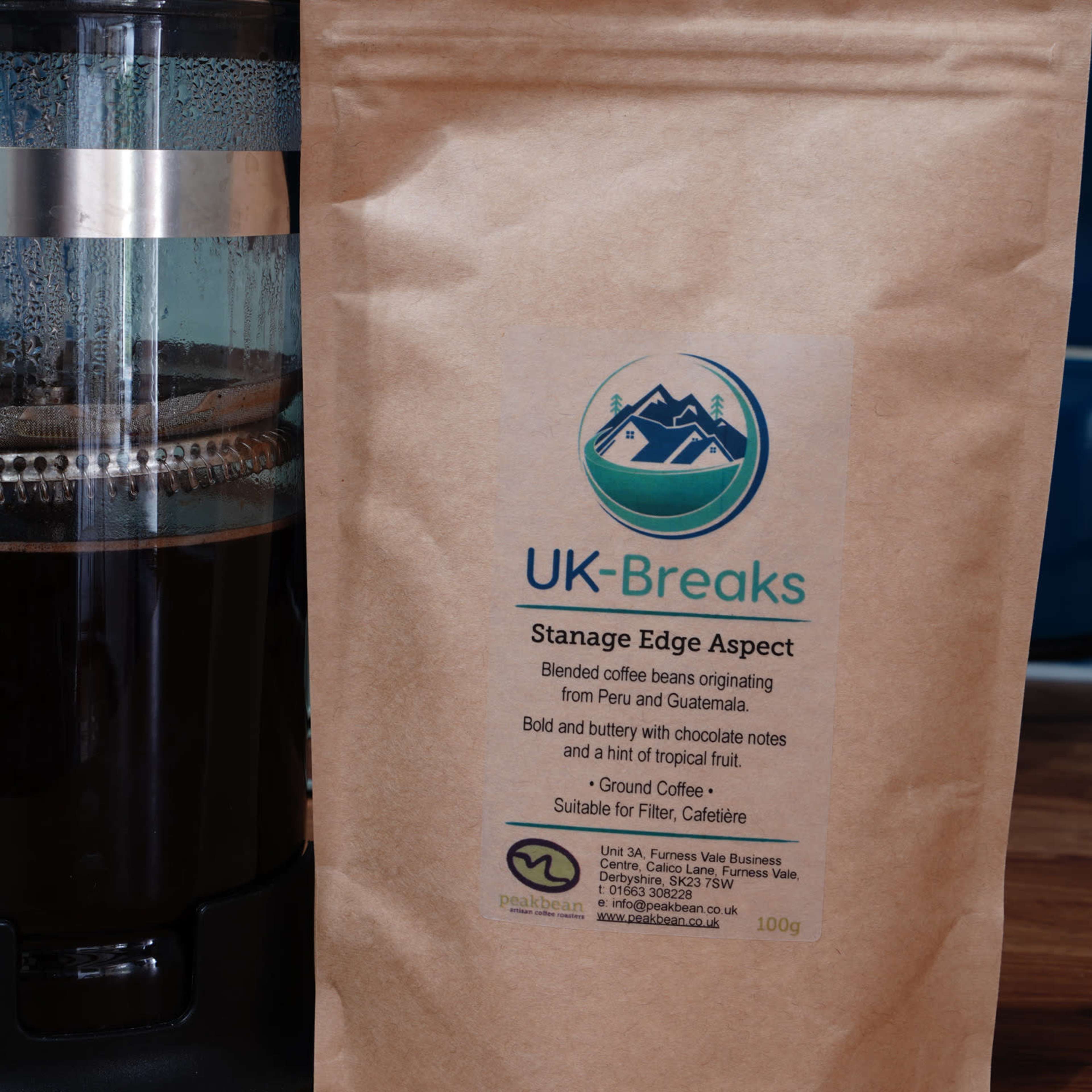A French press filled with brewed coffee stands next to a brown paper bag labeled "UK-Breaks Stanage Edge Aspect."