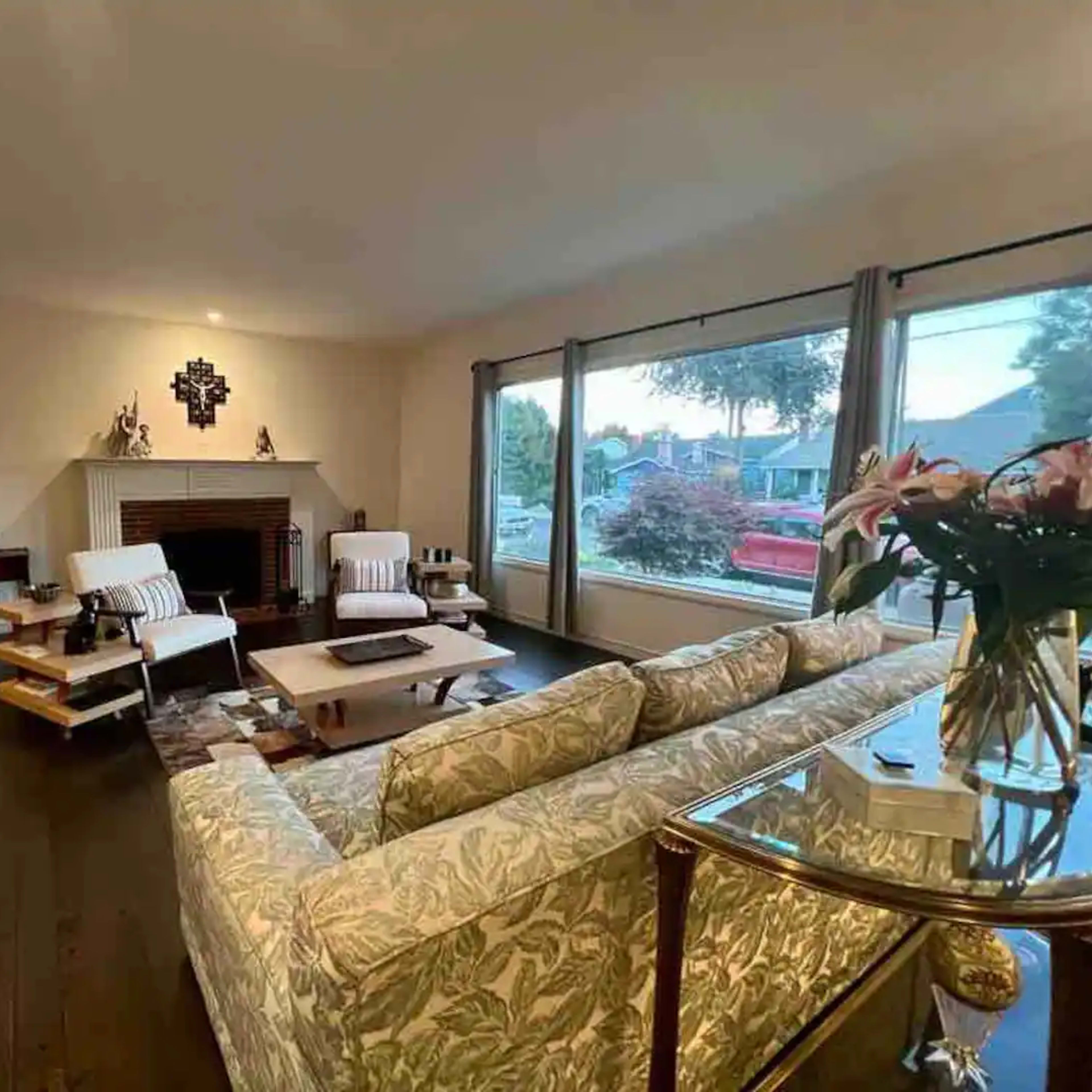 The living room features a floral-patterned sofa, two armchairs, a coffee table, and large windows providing a view of the outside.