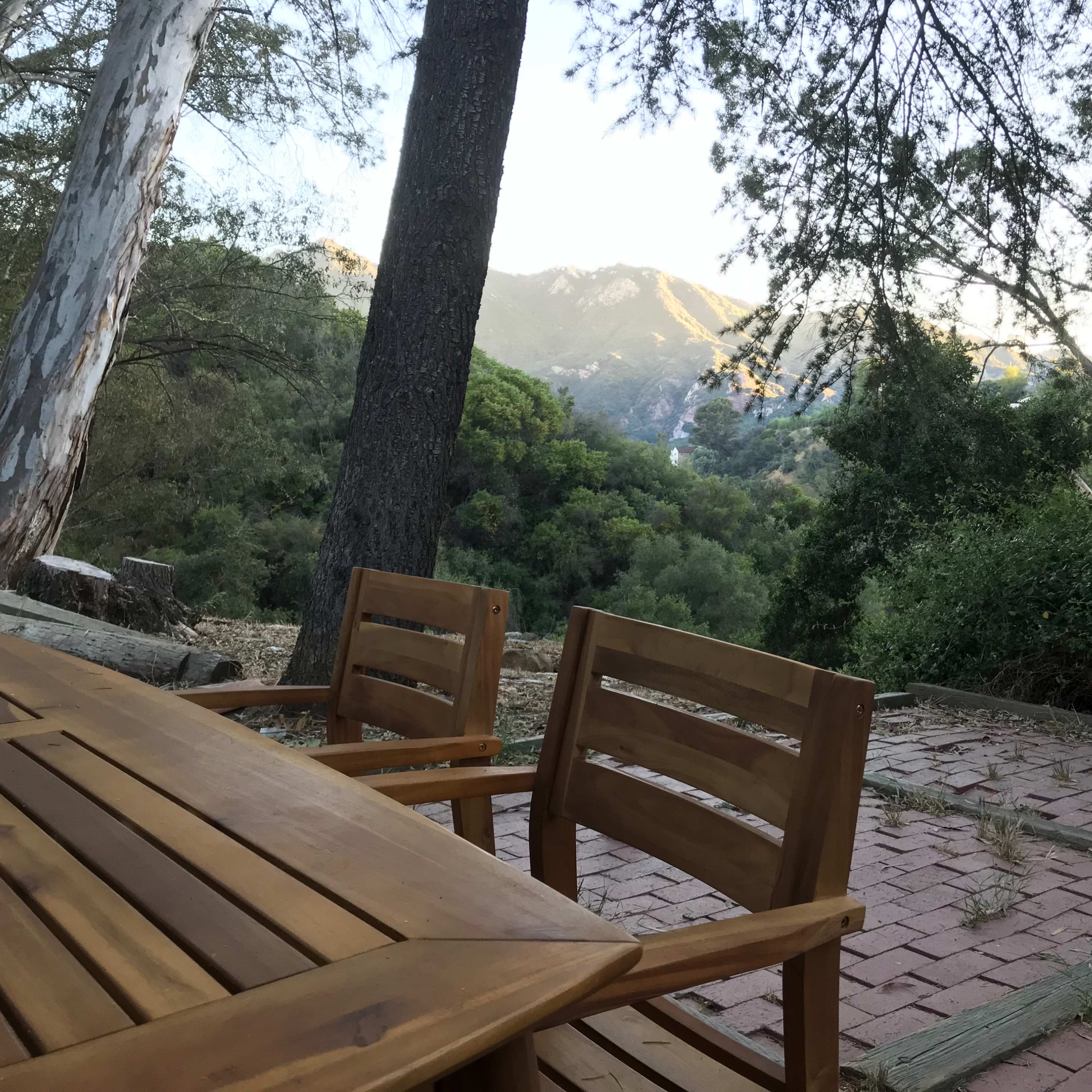A wooden table and chairs are set on a brick patio, facing a view of mountains and trees in the background.