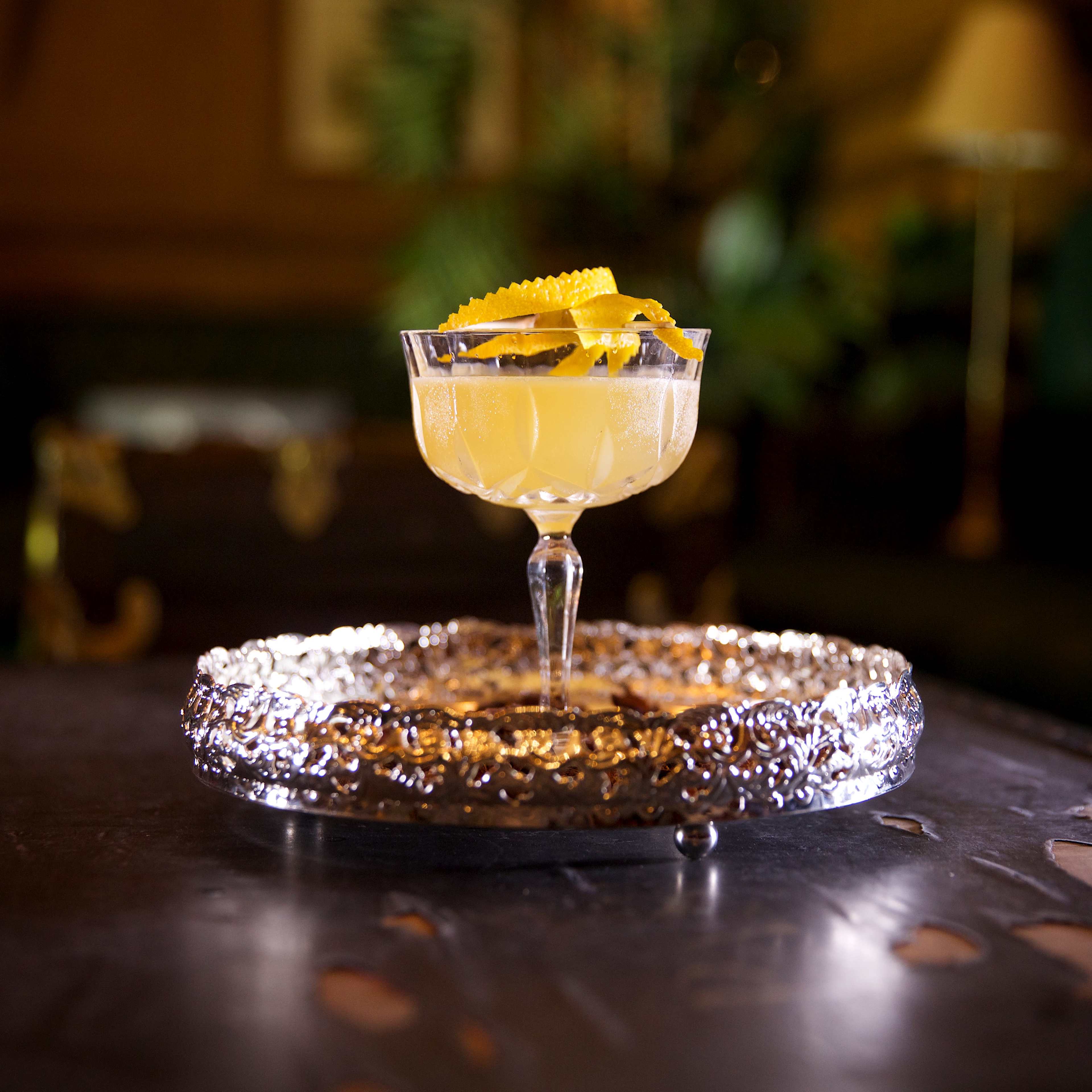 A cocktail served in a vintage glass, garnished with lemon zest, sits on an ornate silver tray.