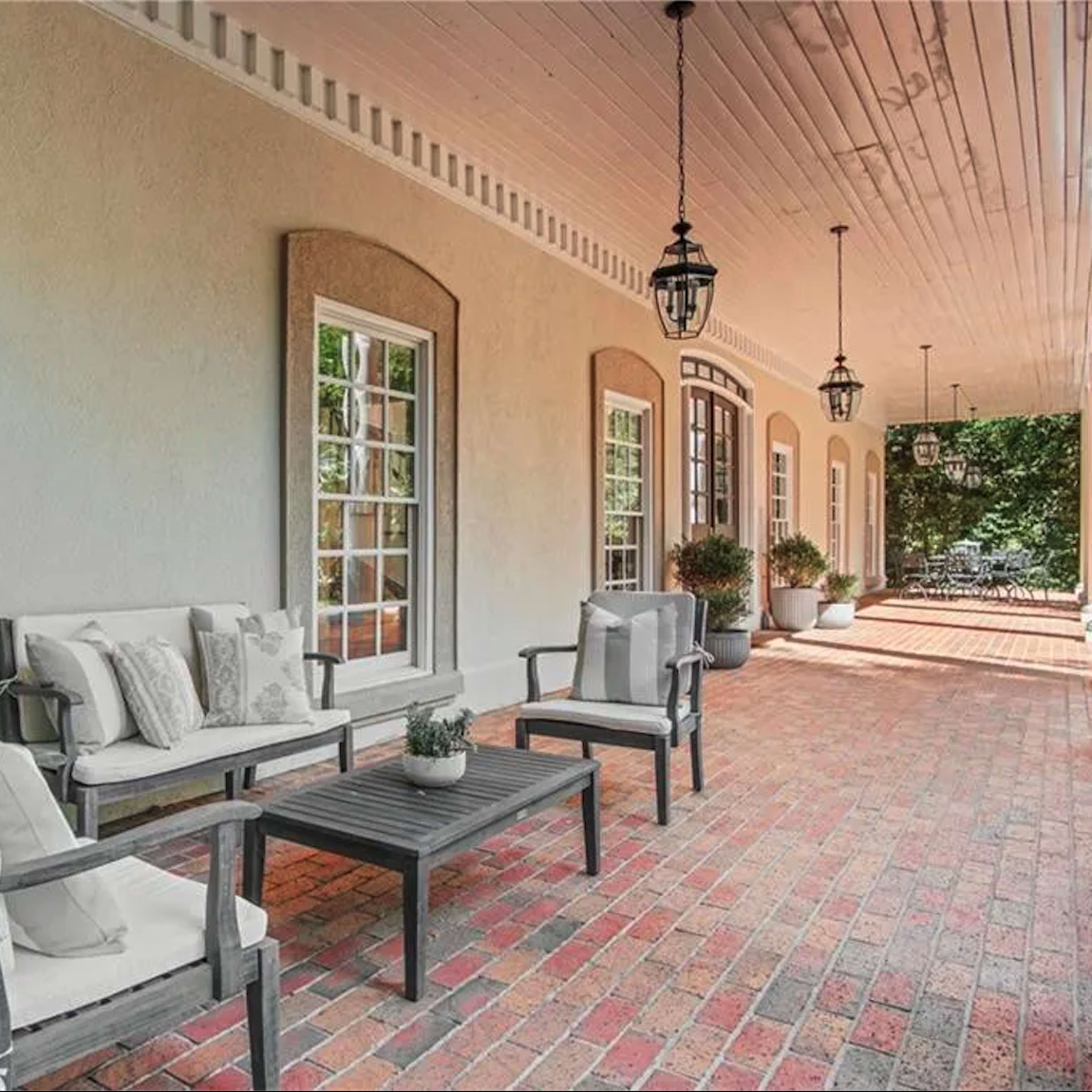 A patio with two chairs and a table on a brick surface, surrounded by large windows and decorative light fixtures hanging from the ceiling.