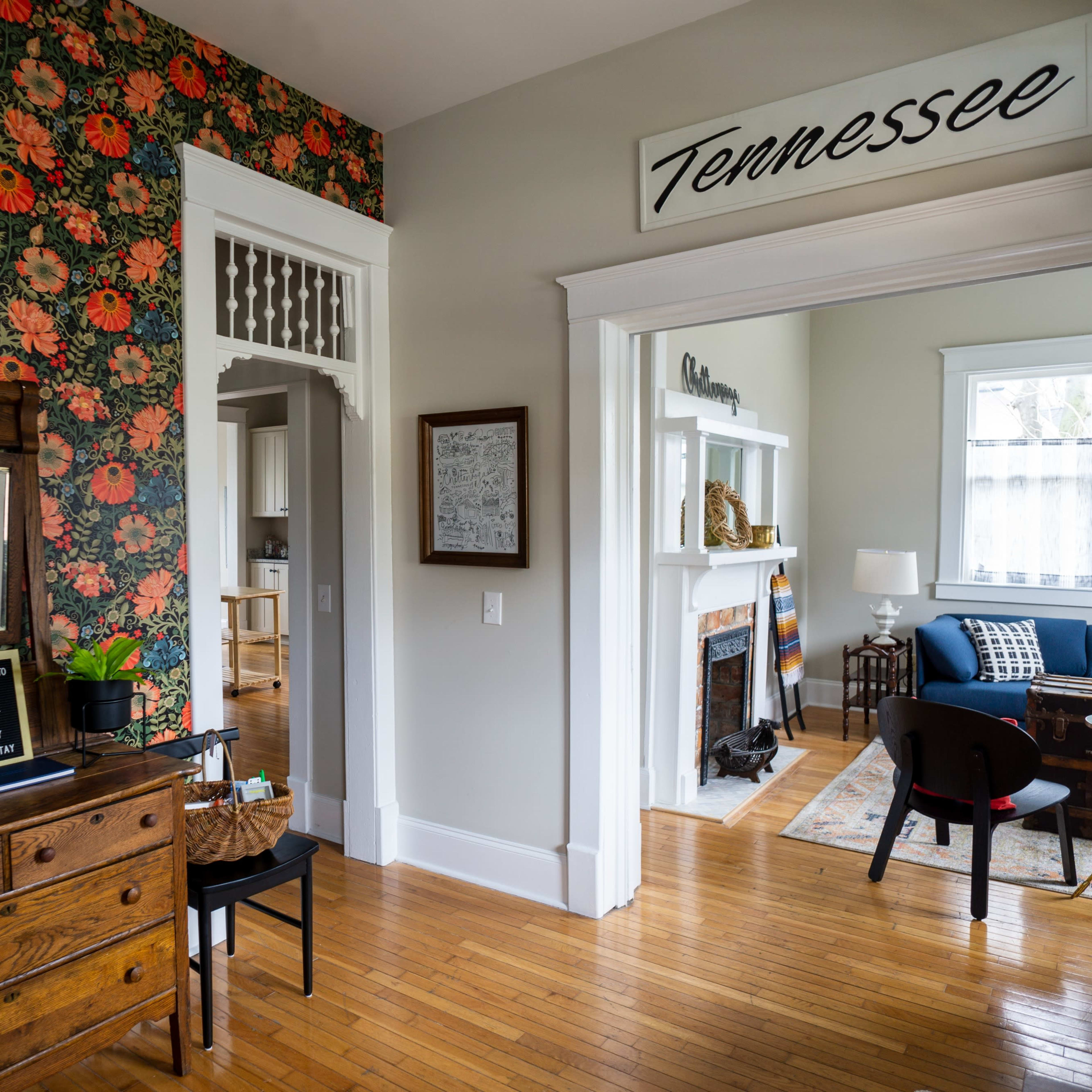 A living space with floral wallpaper, a wooden dresser with a lamp, and a cozy seating area visible through an open doorway.