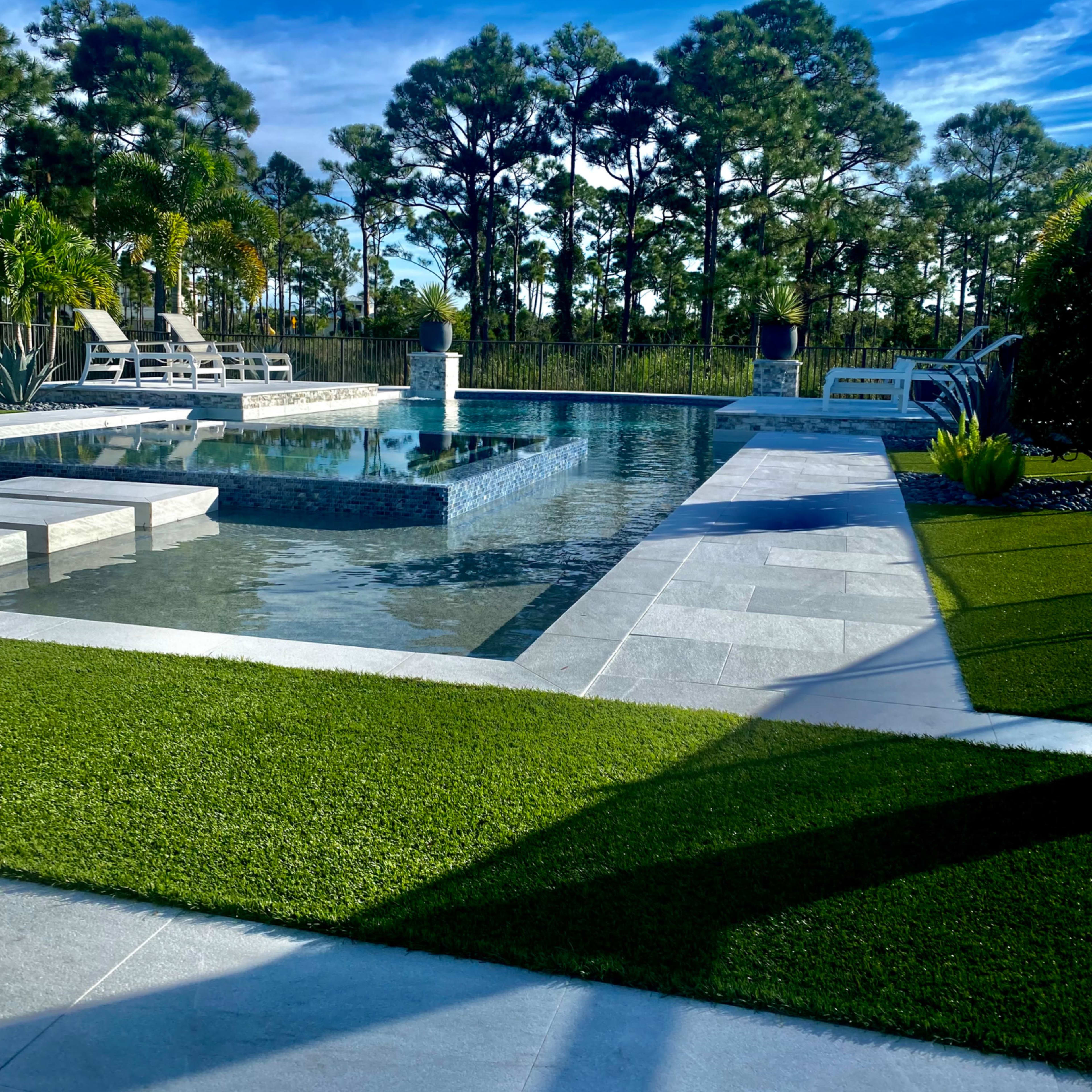 A modern outdoor swimming pool area surrounded by green grass and trees under a blue sky.
