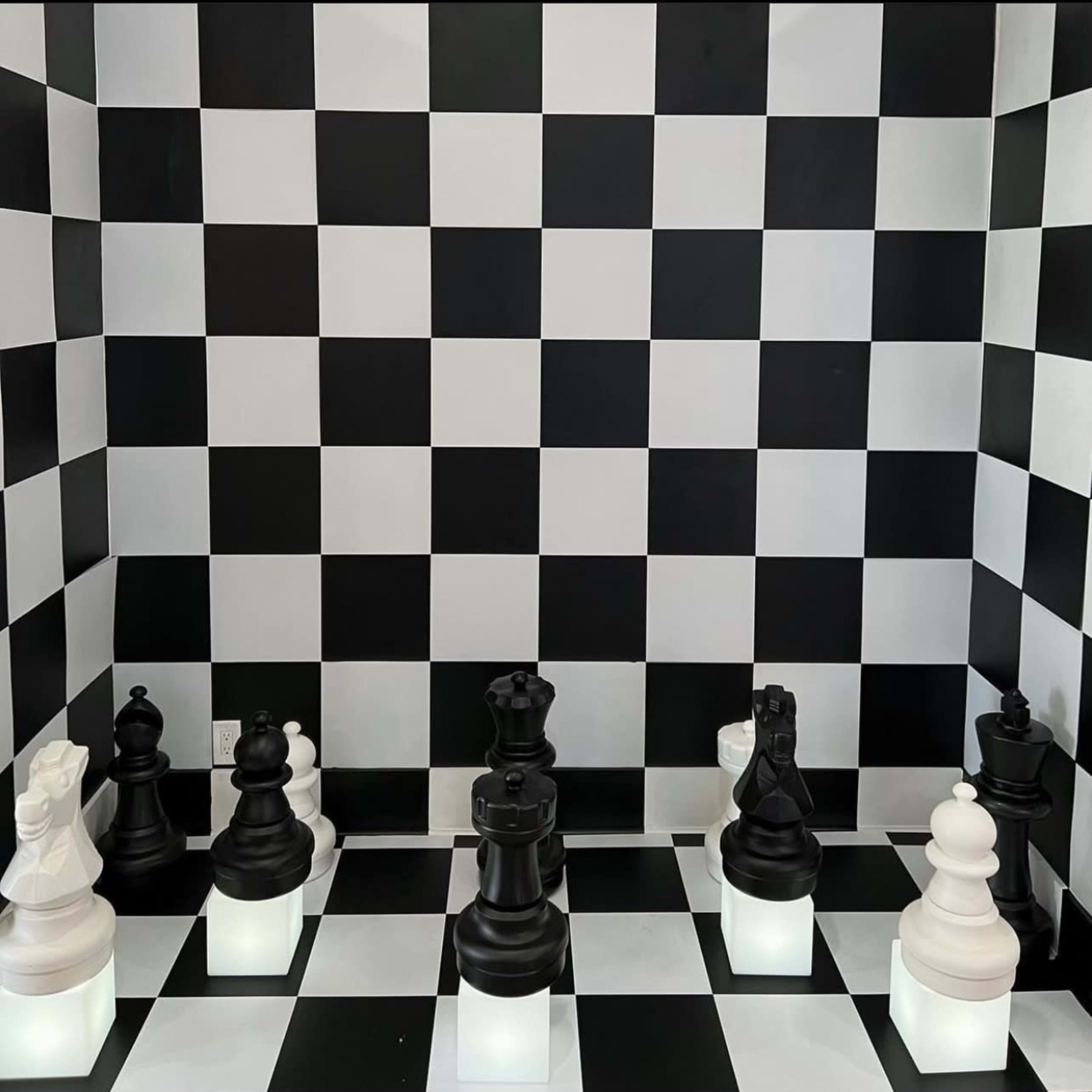 A chess game is set up on a black and white checkered board, featuring large black and white pieces illuminated from below.
