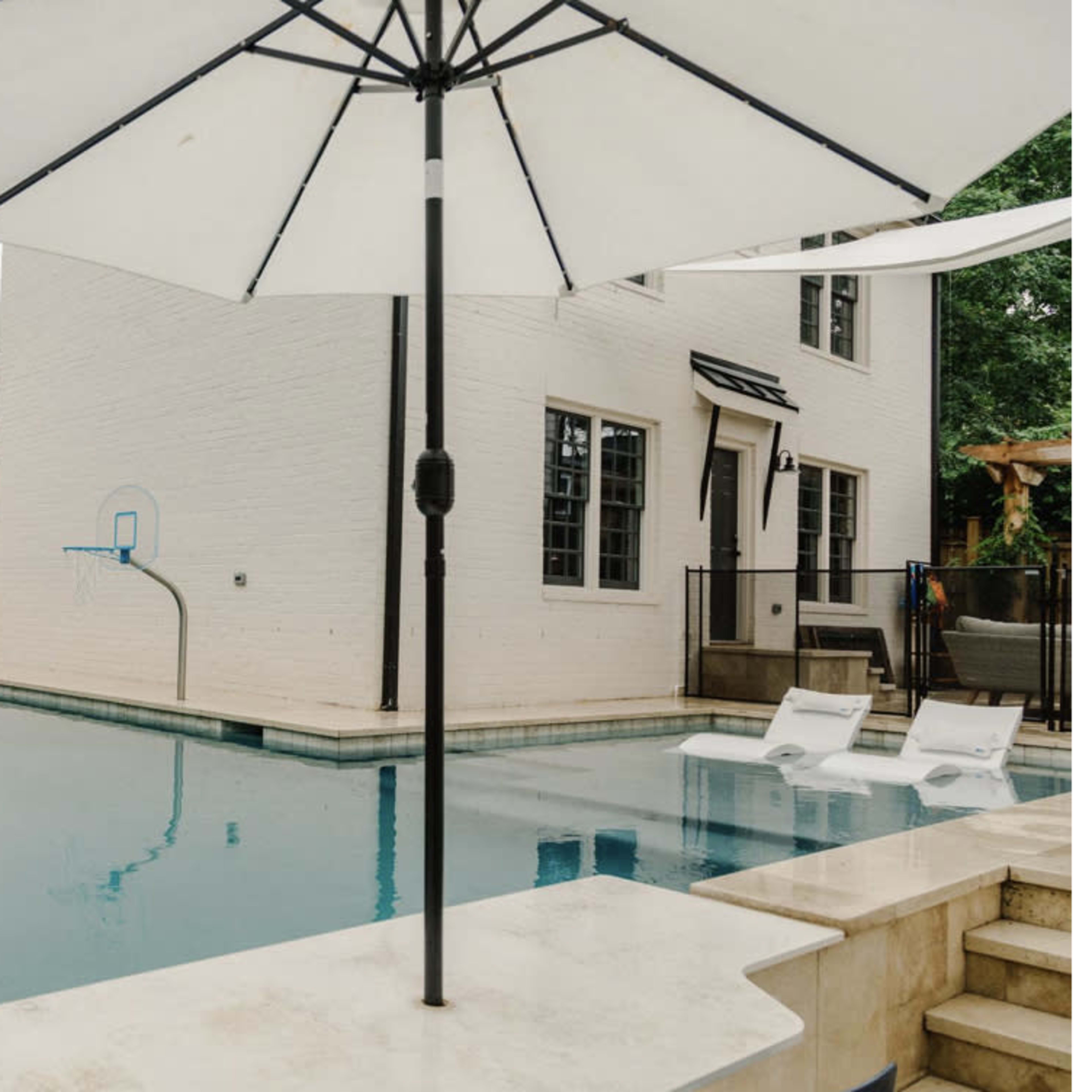A rectangular swimming pool with a slide, lounge chairs, and a large umbrella set in a backyard surrounded by a wooden fence.
