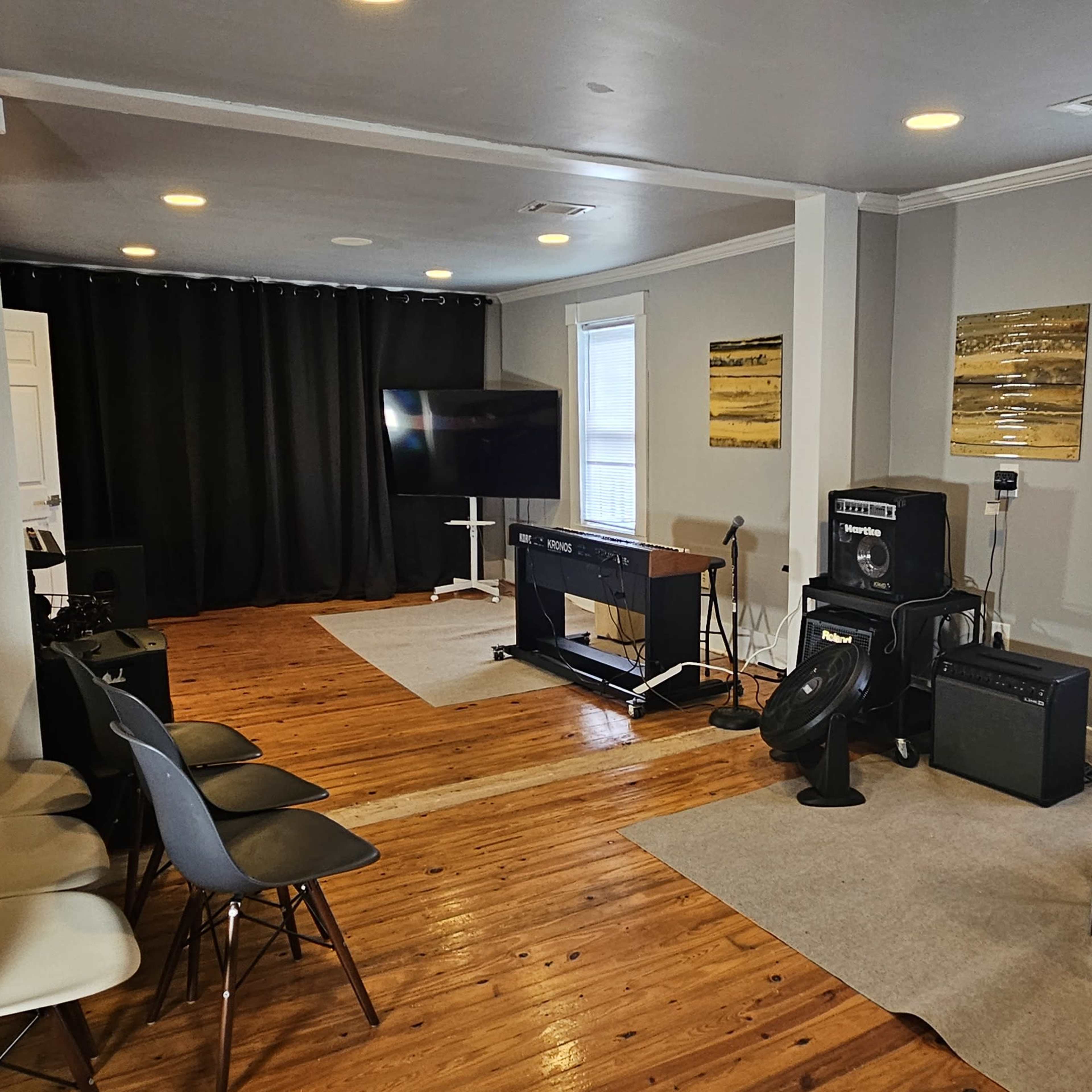The room features a music rehearsal setup with chairs, a television, various instruments, and sound equipment against neutral-colored walls.