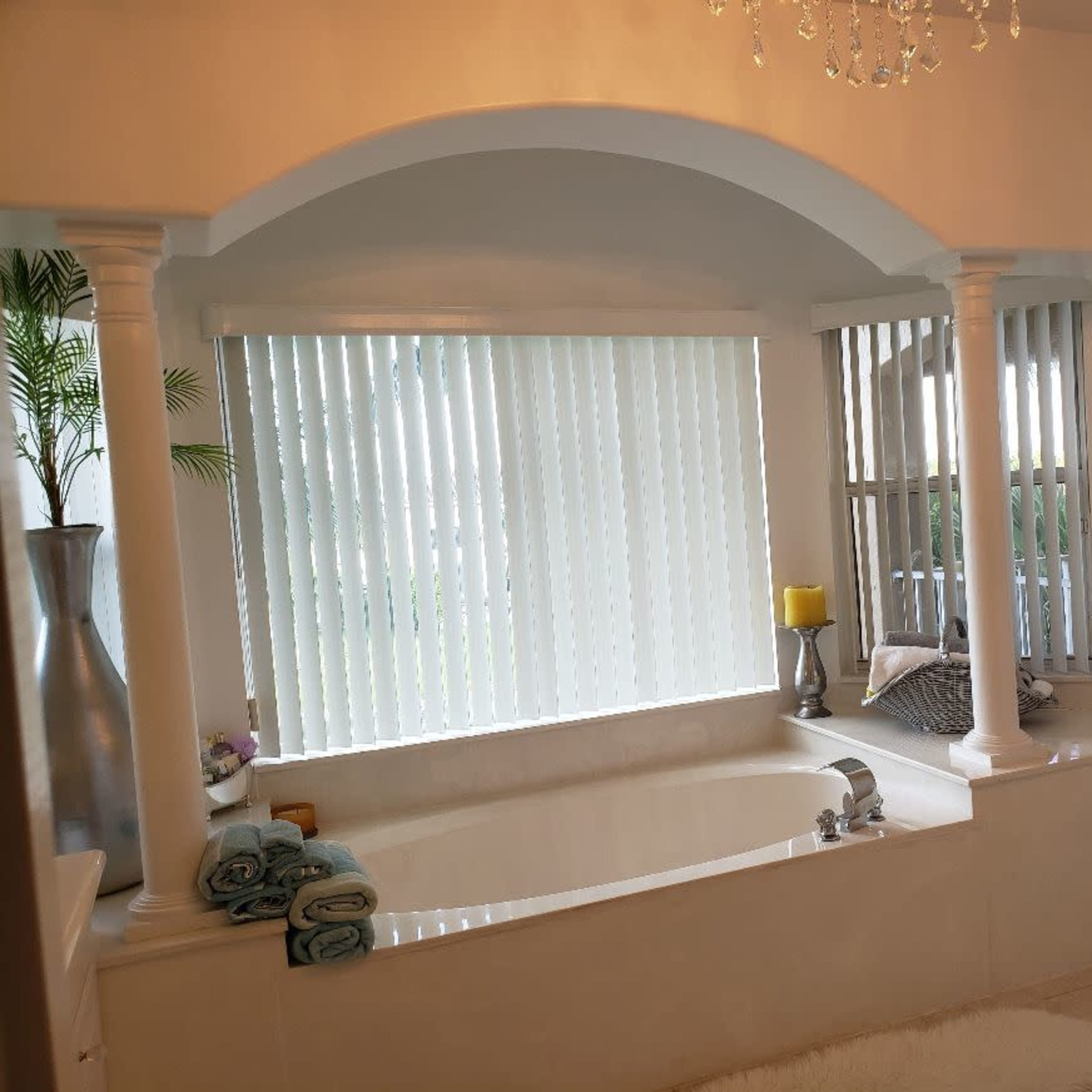 A spacious bathroom featuring a bathtub surrounded by white columns and vertical blinds, with towels neatly arranged nearby.