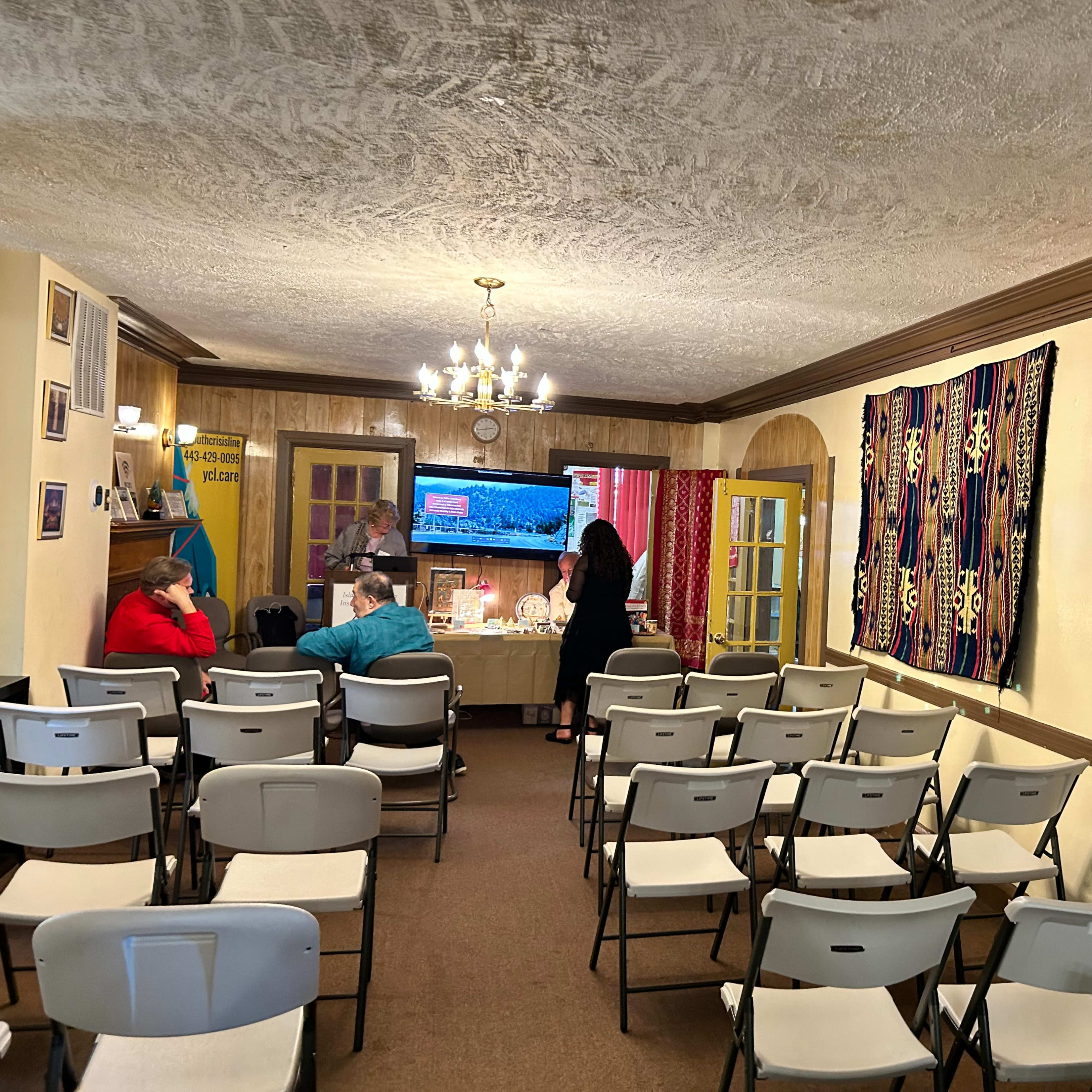 An interior space with folding chairs arranged in rows, a television screen on the wall, and colorful woven textiles hanging beside an open door.