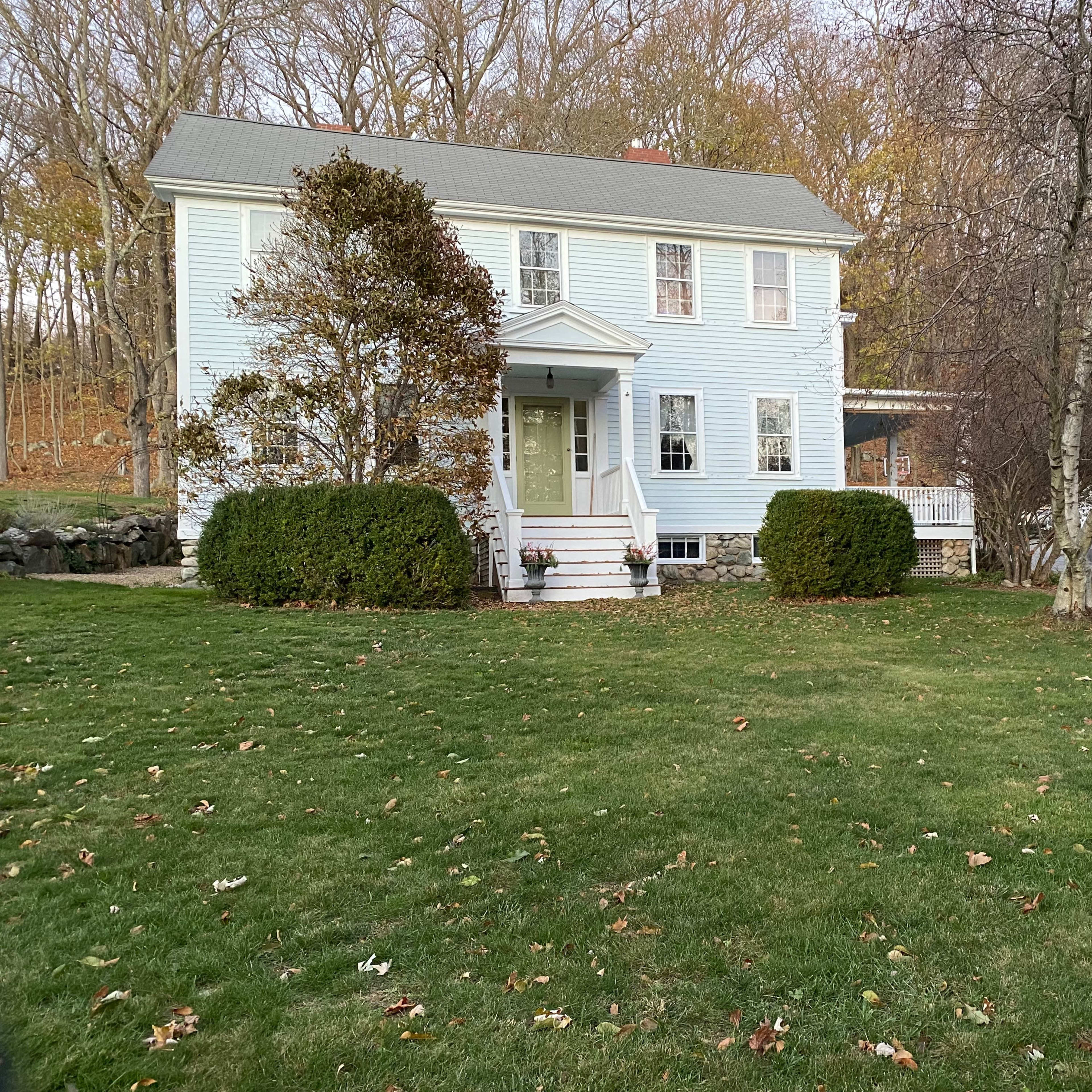 A light blue two-story house with a front porch is set on a grassy lawn surrounded by trees in autumn.