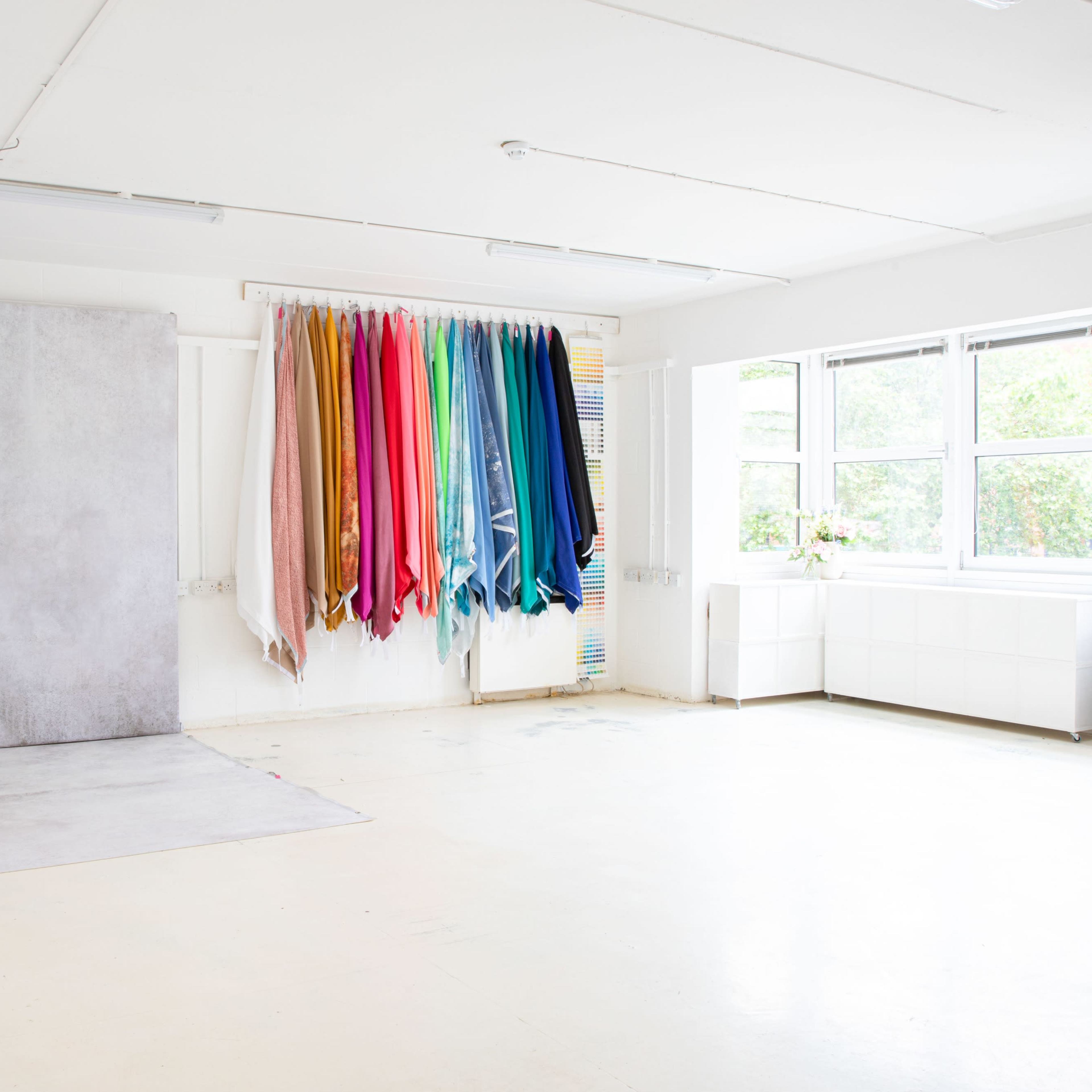 A bright studio space with a gray backdrop and a vertical display of colorful fabric rolls hanging on the wall.