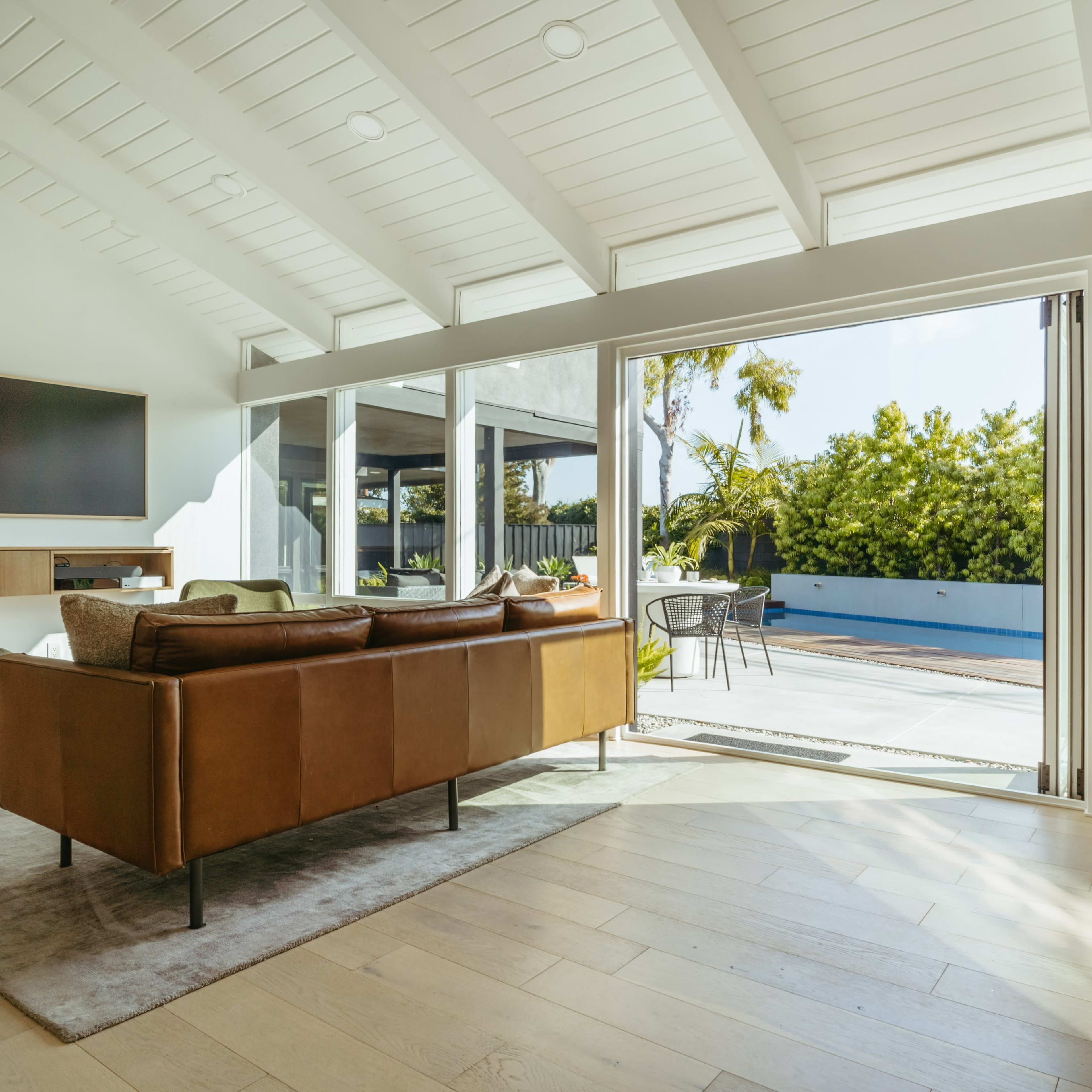 A modern living room features a brown leather sofa, a flat-screen TV on the wall, and large glass doors that open to a patio with outdoor seating and greenery.