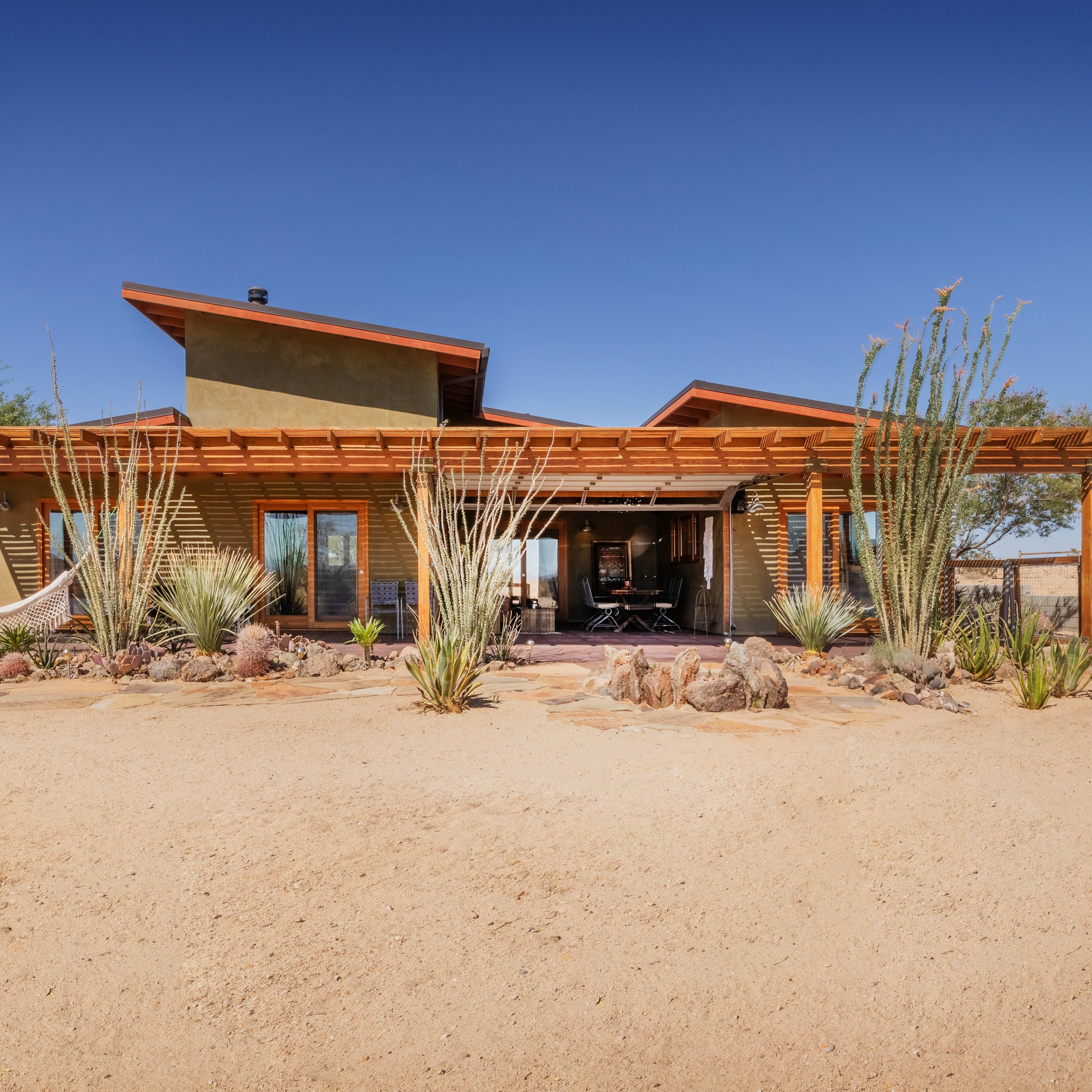 A modern desert house with wooden accents and large windows is surrounded by cacti and gravel landscaping.