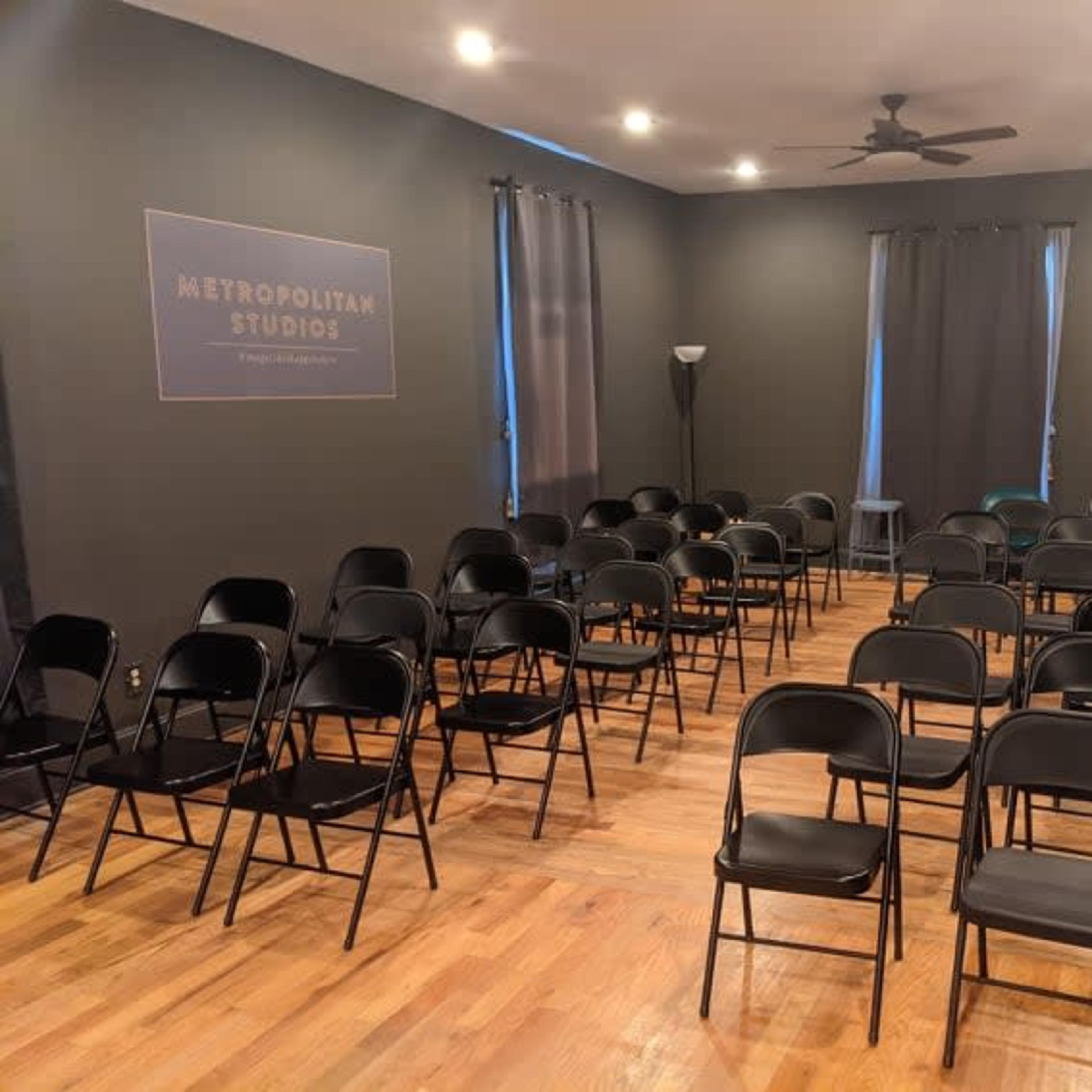 The room is set up with rows of black folding chairs on a wooden floor, featuring dark walls and windows covered by curtains.