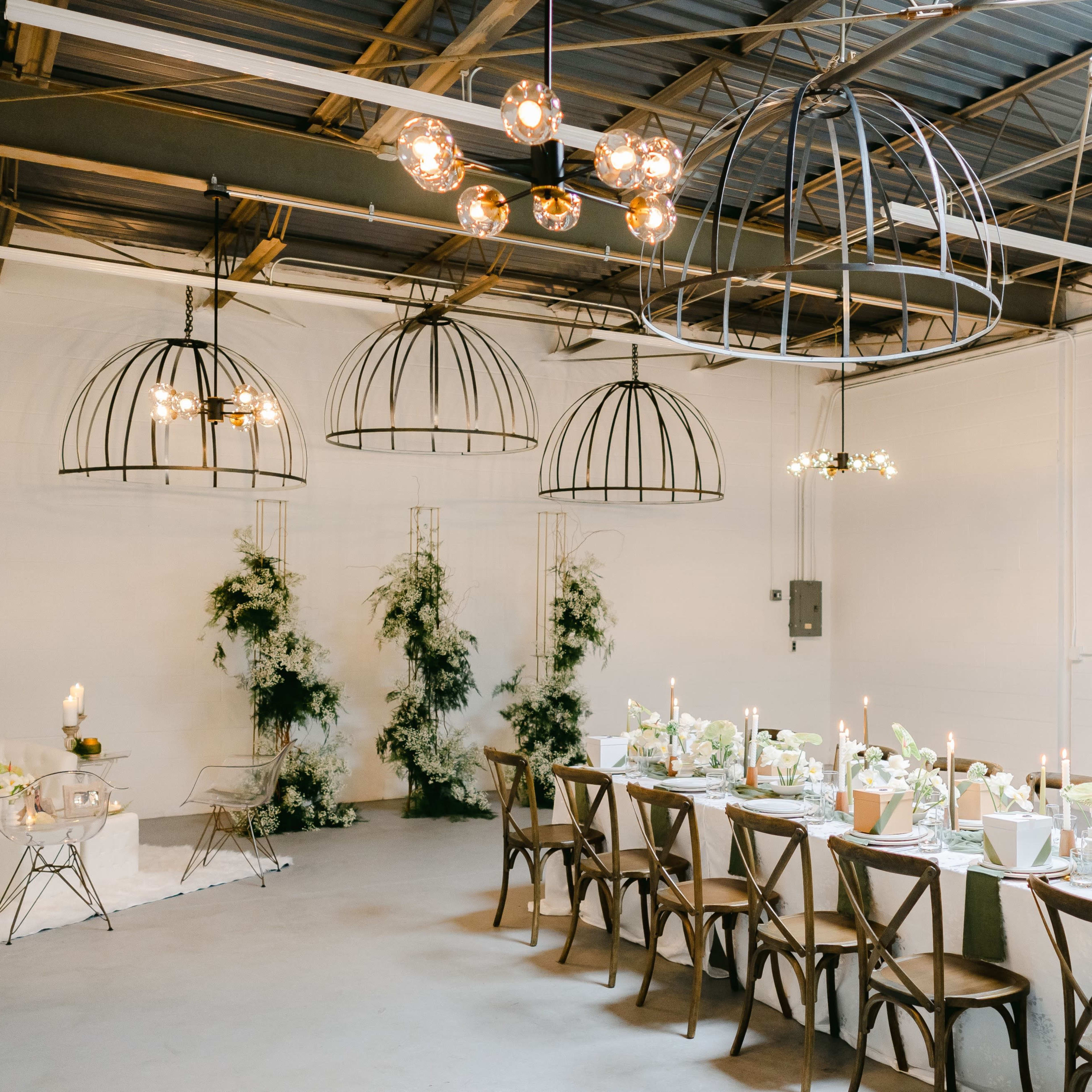 A dining area features a long table set with floral arrangements and candles, surrounded by industrial-style light fixtures and greenery accents.