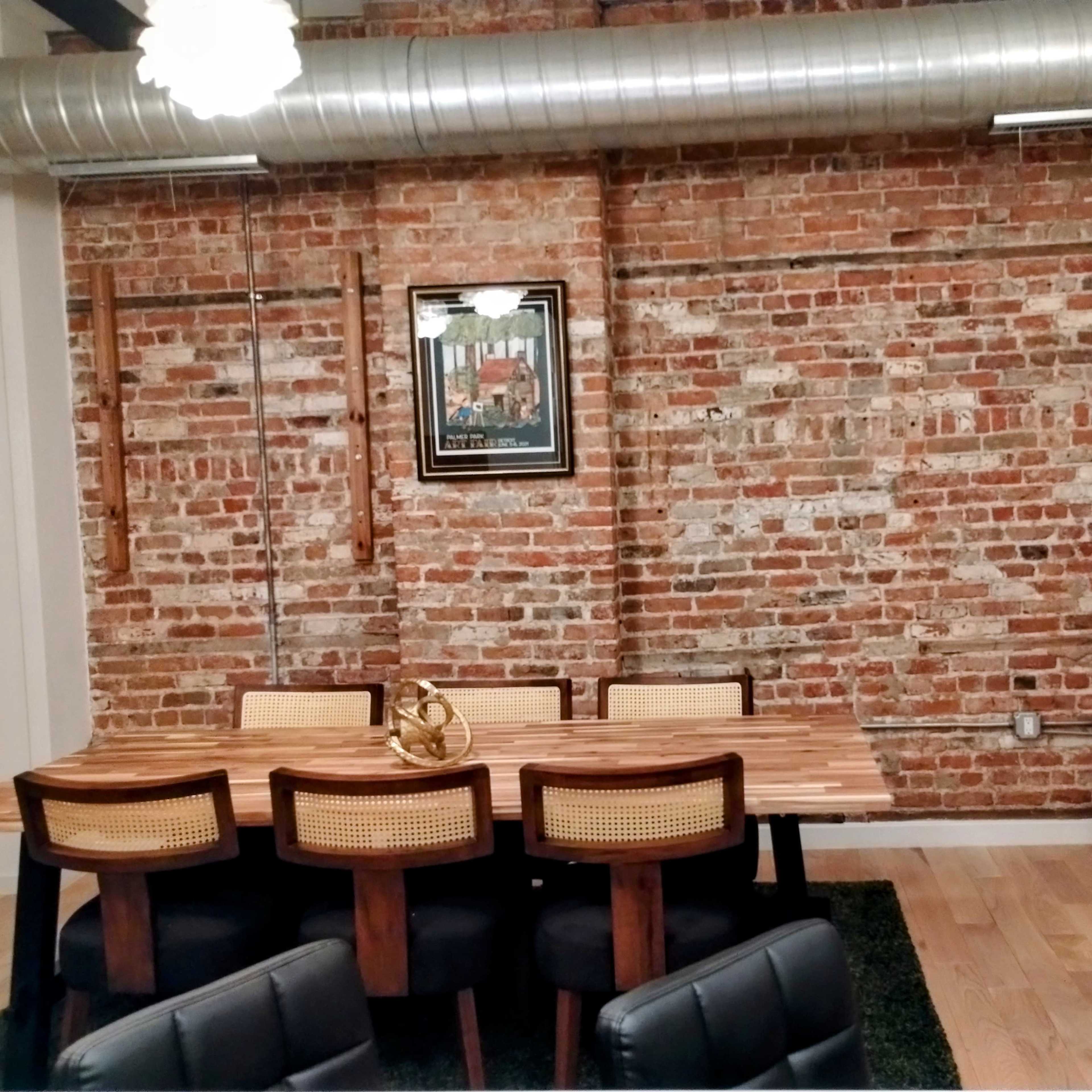 A dining area with a wooden table and chairs against a backdrop of exposed brick walls, featuring a decorative framed painting and a wicker chair in the corner.
