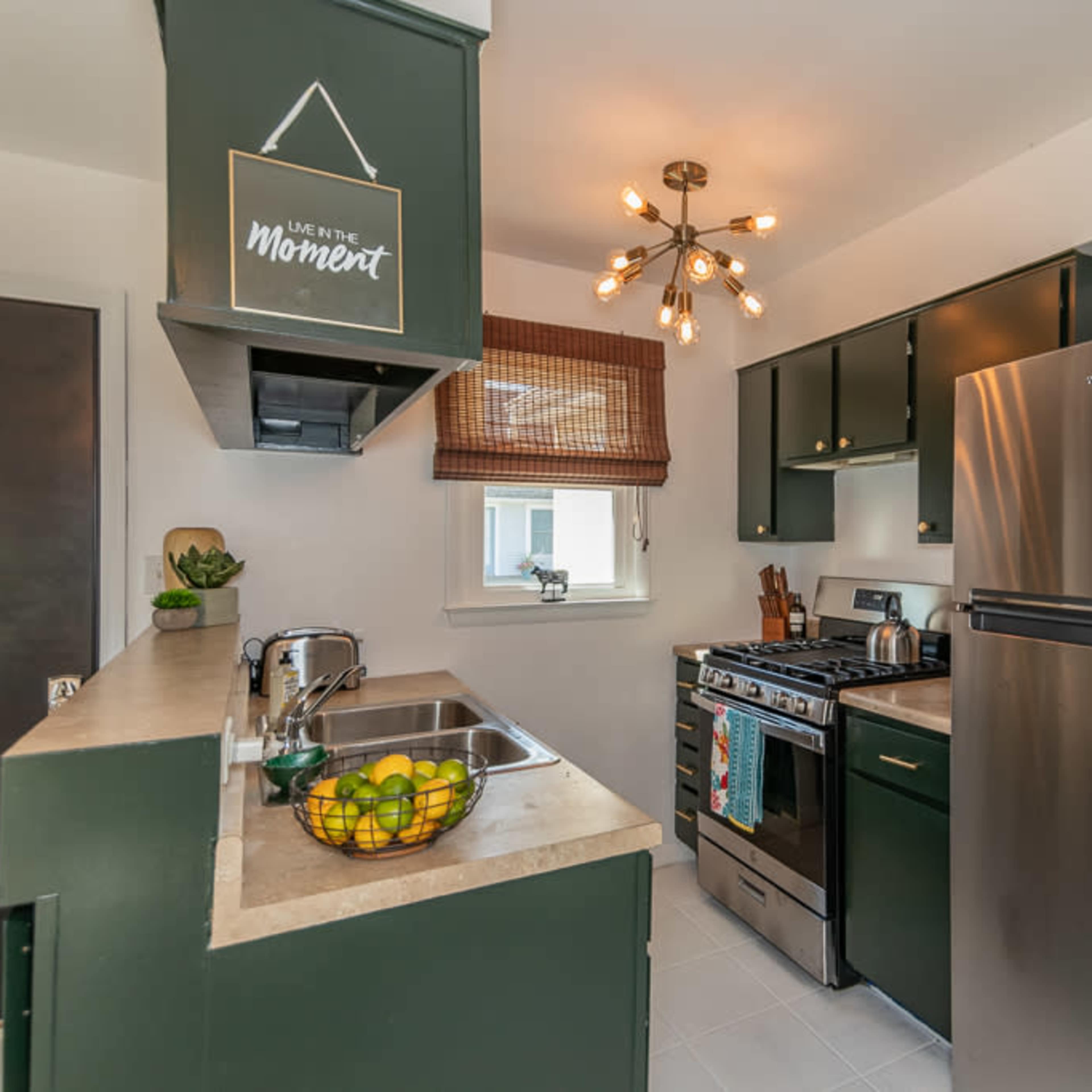 The kitchen features dark green cabinetry, stainless steel appliances, and a central light fixture, with a small window and a bowl of lemons on the counter.