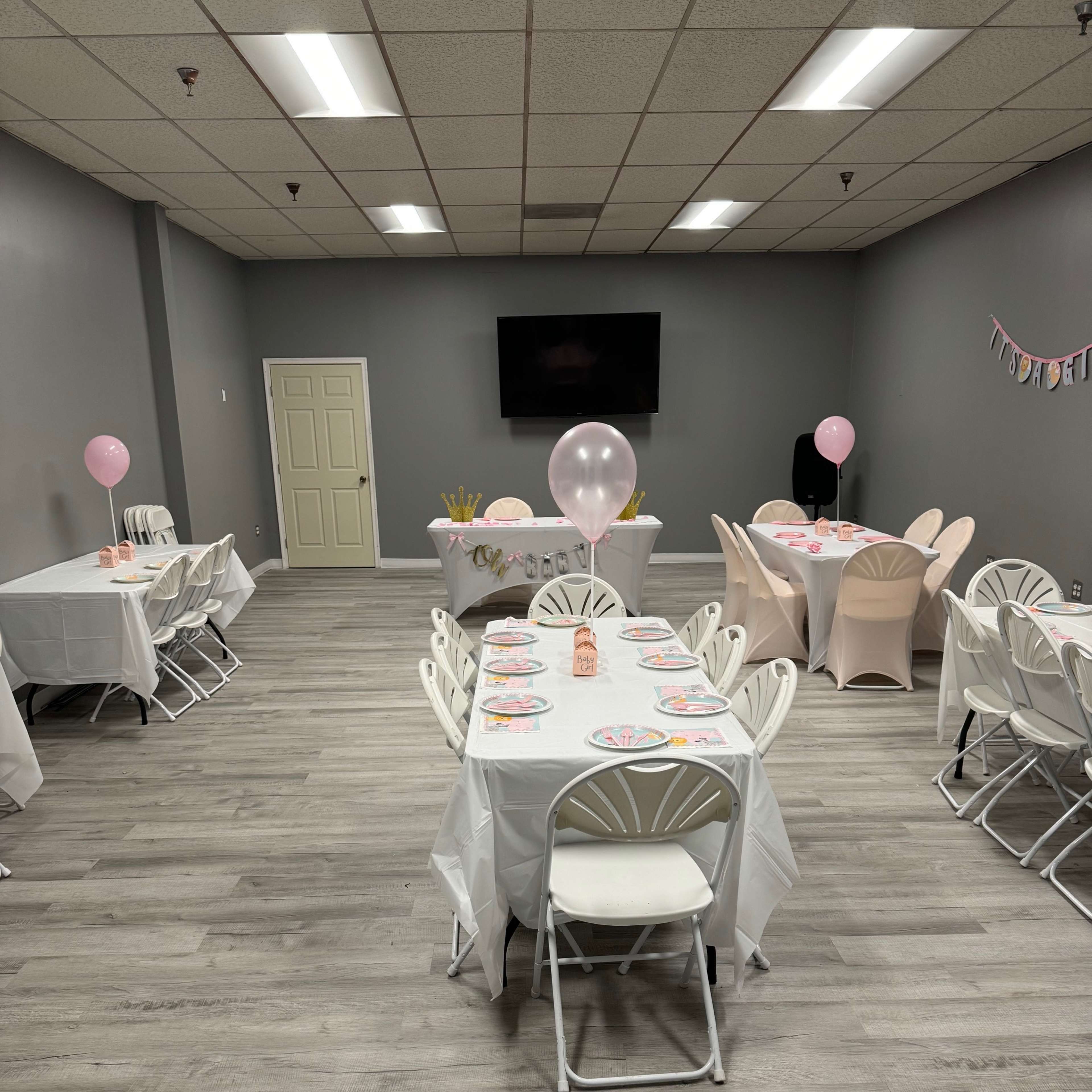 A room set up for a party, featuring several tables with white tablecloths and pink decorations, along with balloons and a television on the wall.