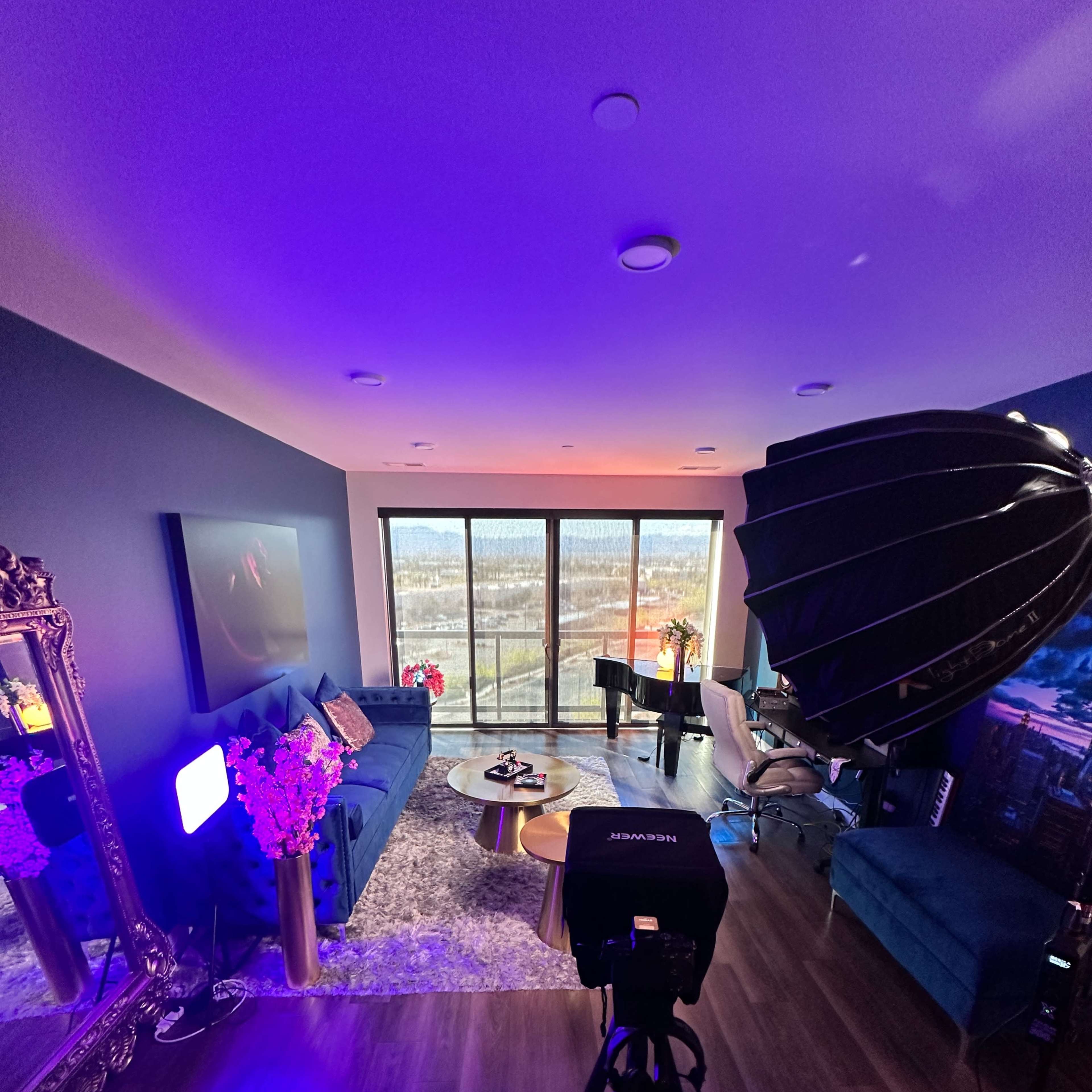 A modern living room features a blue sofa, a piano, and large windows overlooking a landscape, with studio lights and a camera set up for a photo shoot.